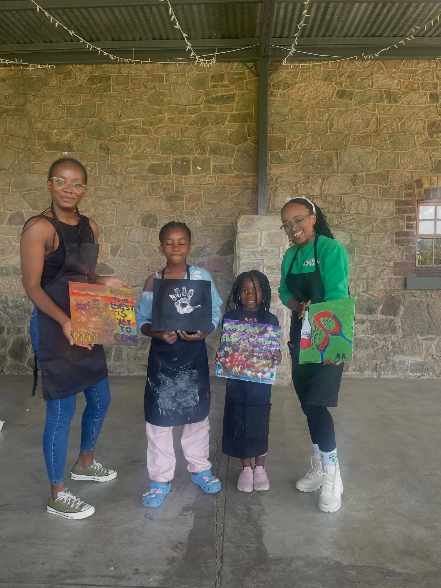 Four women and two children standing in front of a brick wall, holding colorful paintings they created, with smiles and relaxed posture.