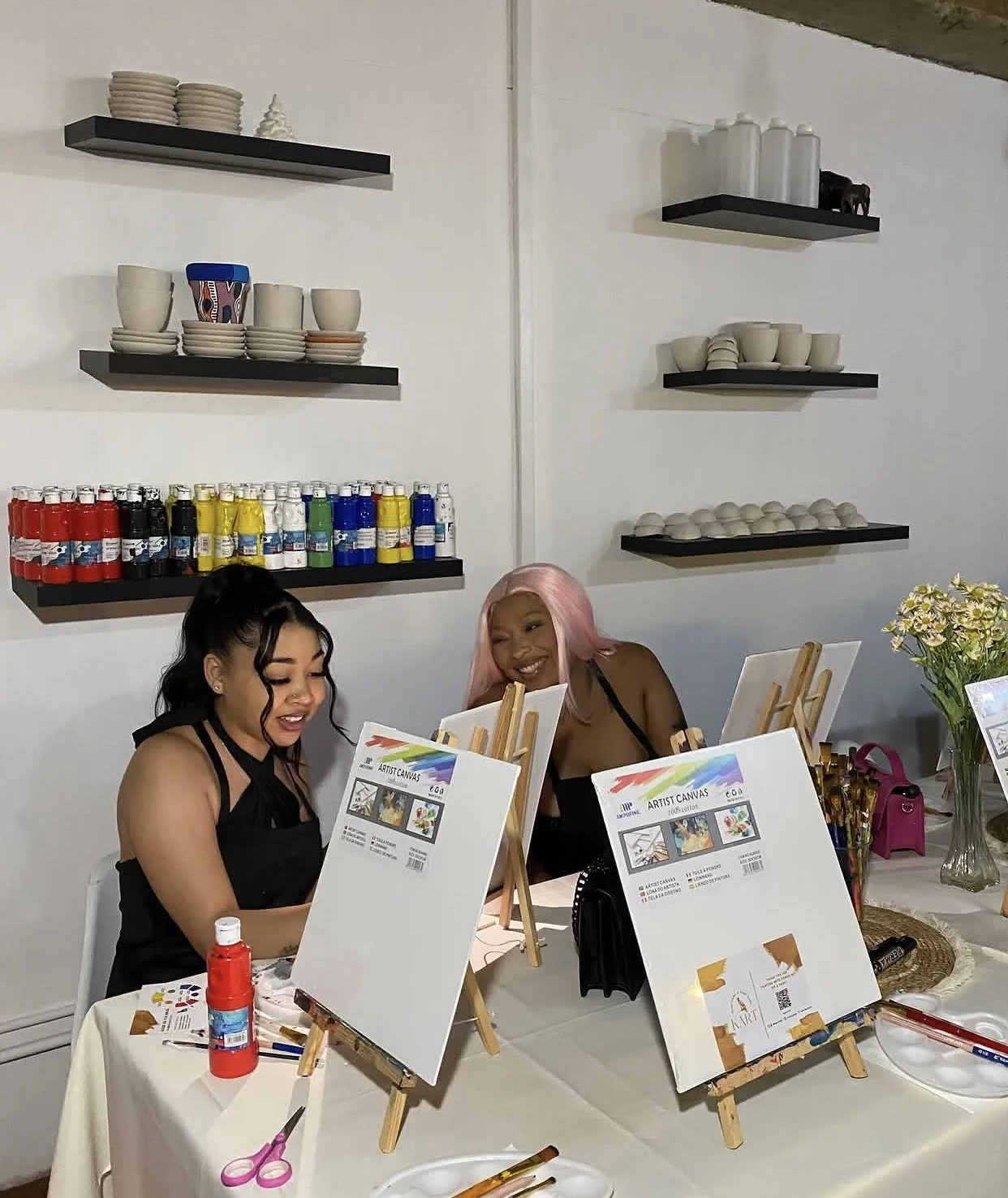 Two women sitting at a table, participating in a painting activity with canvases on small easels. They are smiling and engaged. The table has painting supplies like scissors, glue, and paint bottles. Behind them is a wall with shelves holding plates, bowls, and decorative items, and a bouquet of flowers is on the side of the table.