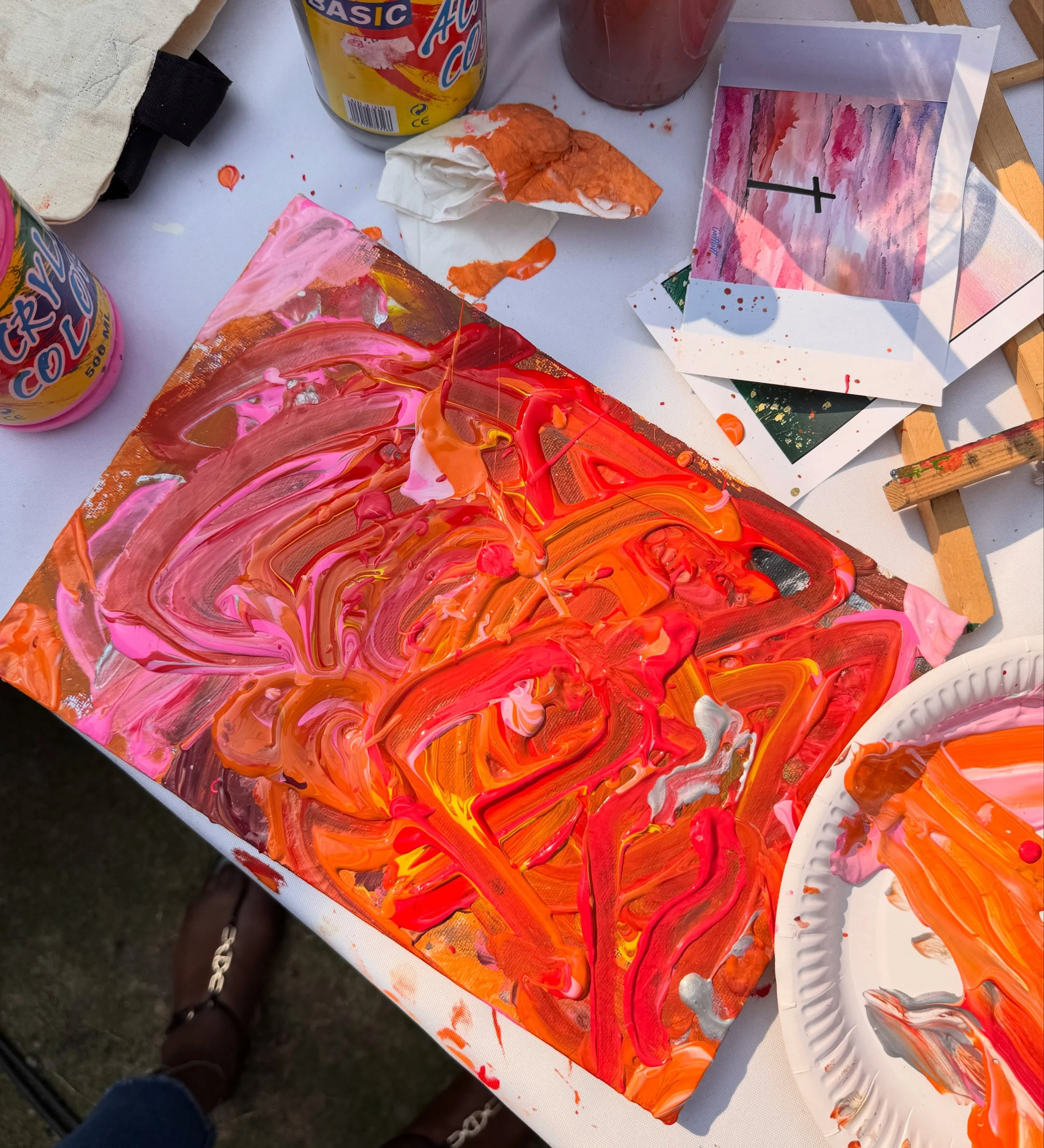 Abstract painting with vibrant orange, pink, and yellow swirls on a canvas, surrounded by paint splatters and two finished paintings in similar color palettes, on a white table.