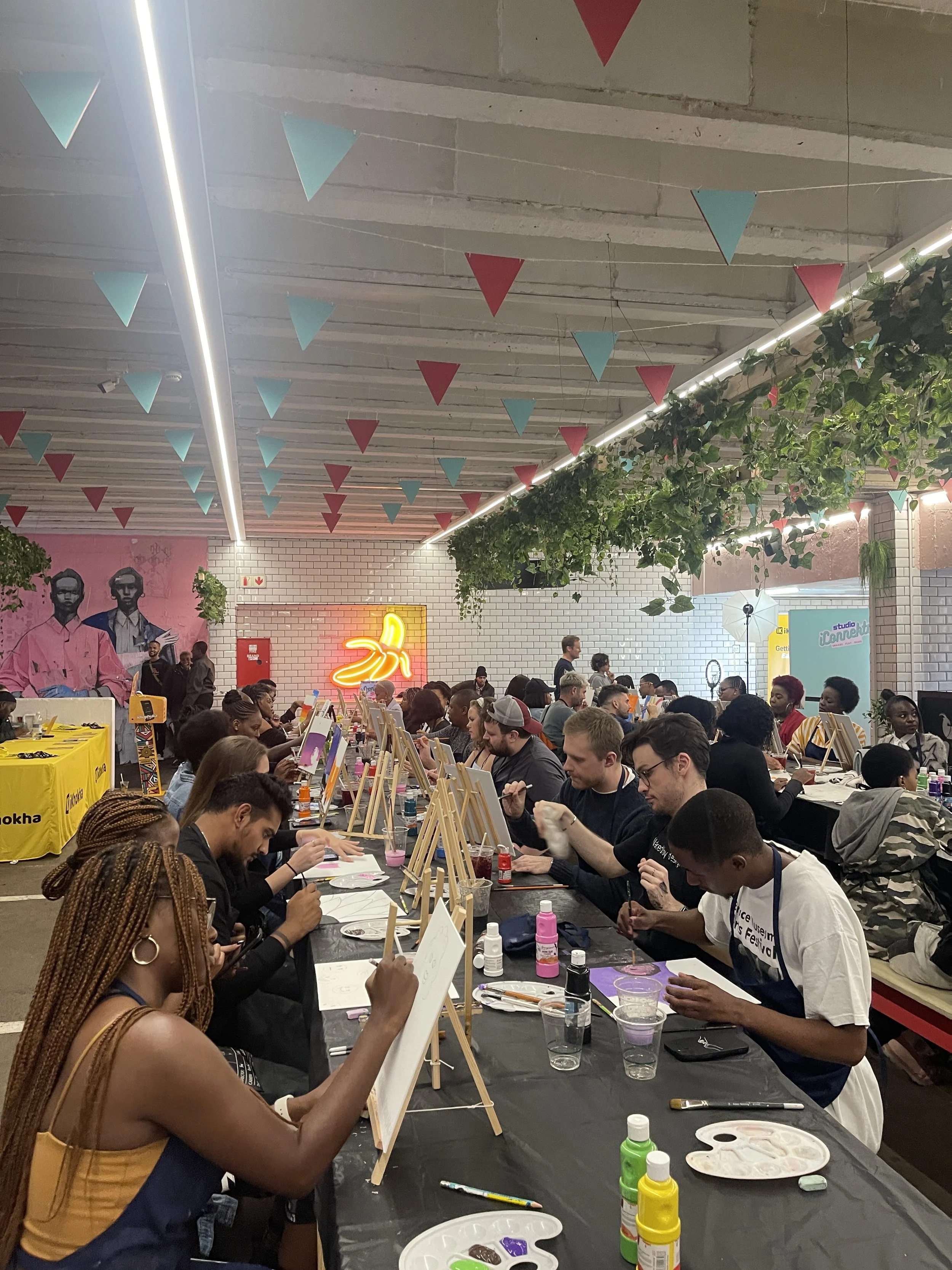 People participating in a group painting activity, sitting at a long table with painting supplies, surrounded by hanging triangular banners and greenery in a decorated indoor space.