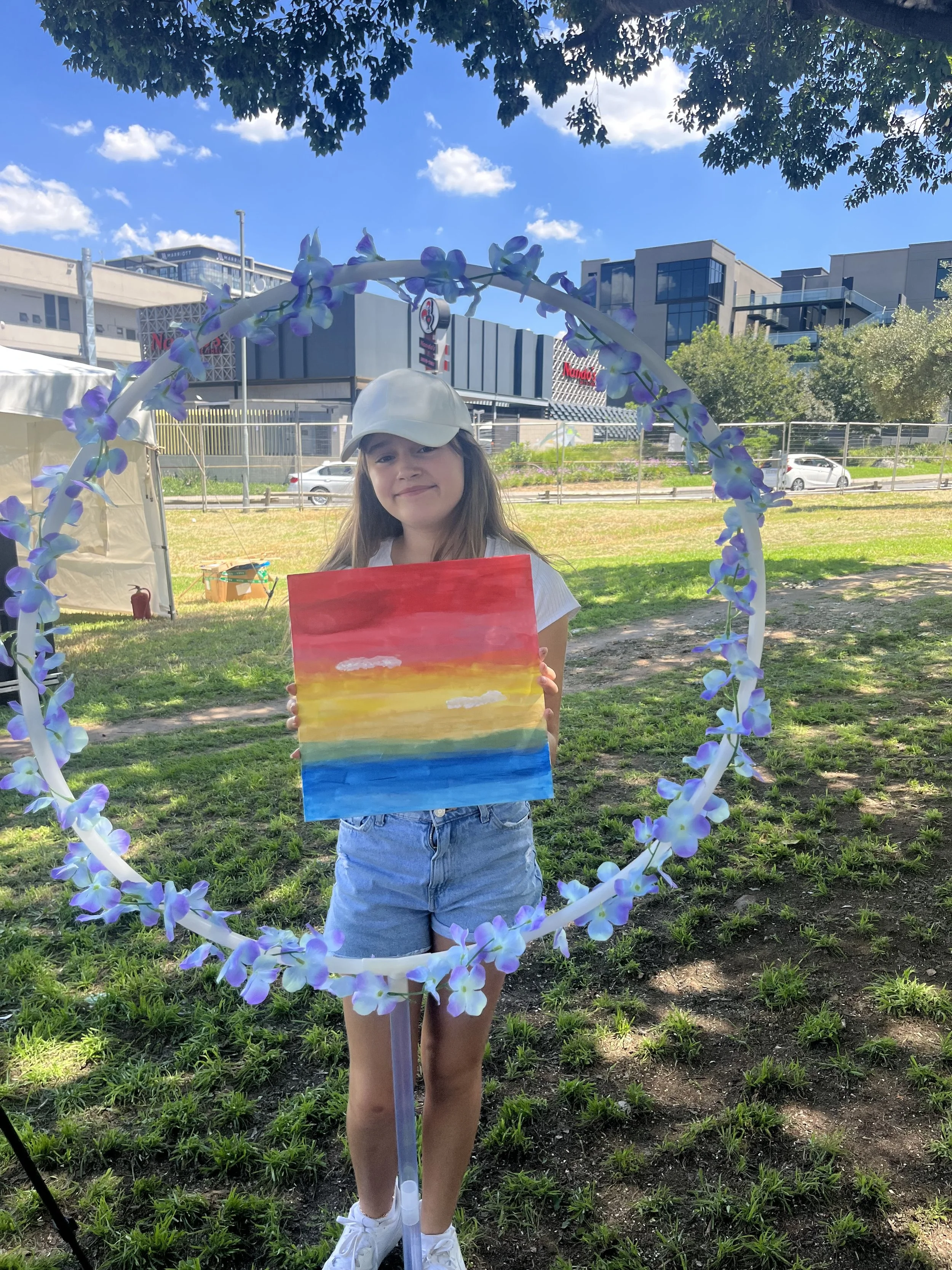 A girl standing outdoors under trees, holding a colorful painting of a sunset over the ocean. She is smiling, wearing a white cap, a short-sleeved white shirt, and denim shorts. She is framed by a decorative circle made of purple and blue flowers.
