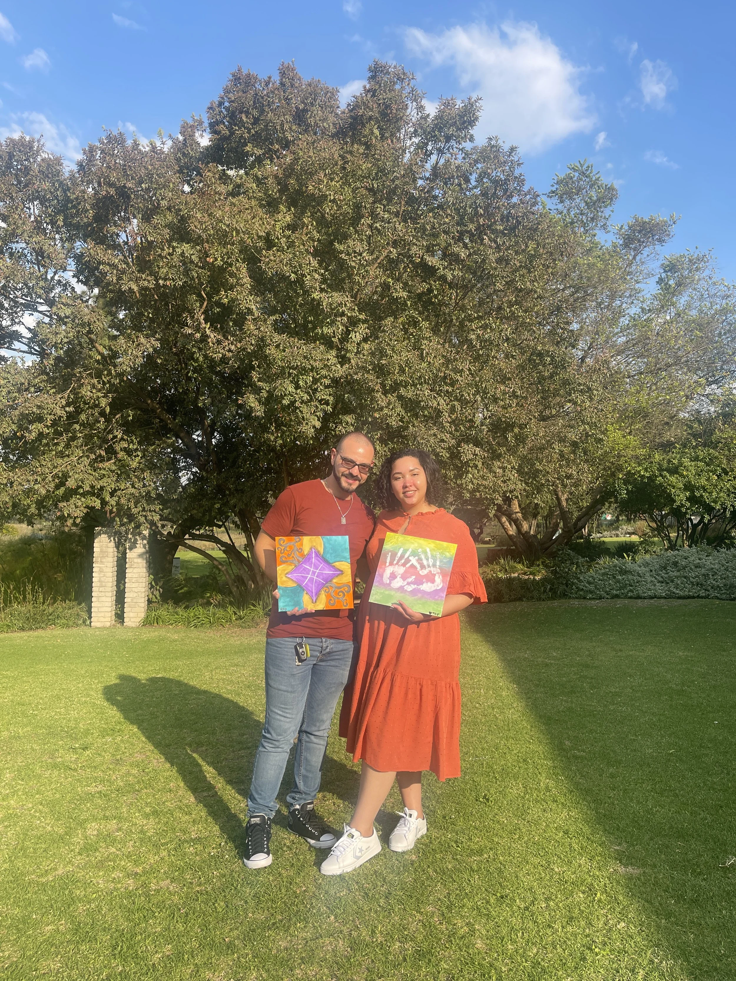 A man and a woman standing on grass in a park, holding colorful paintings, with a large tree and blue sky in the background.