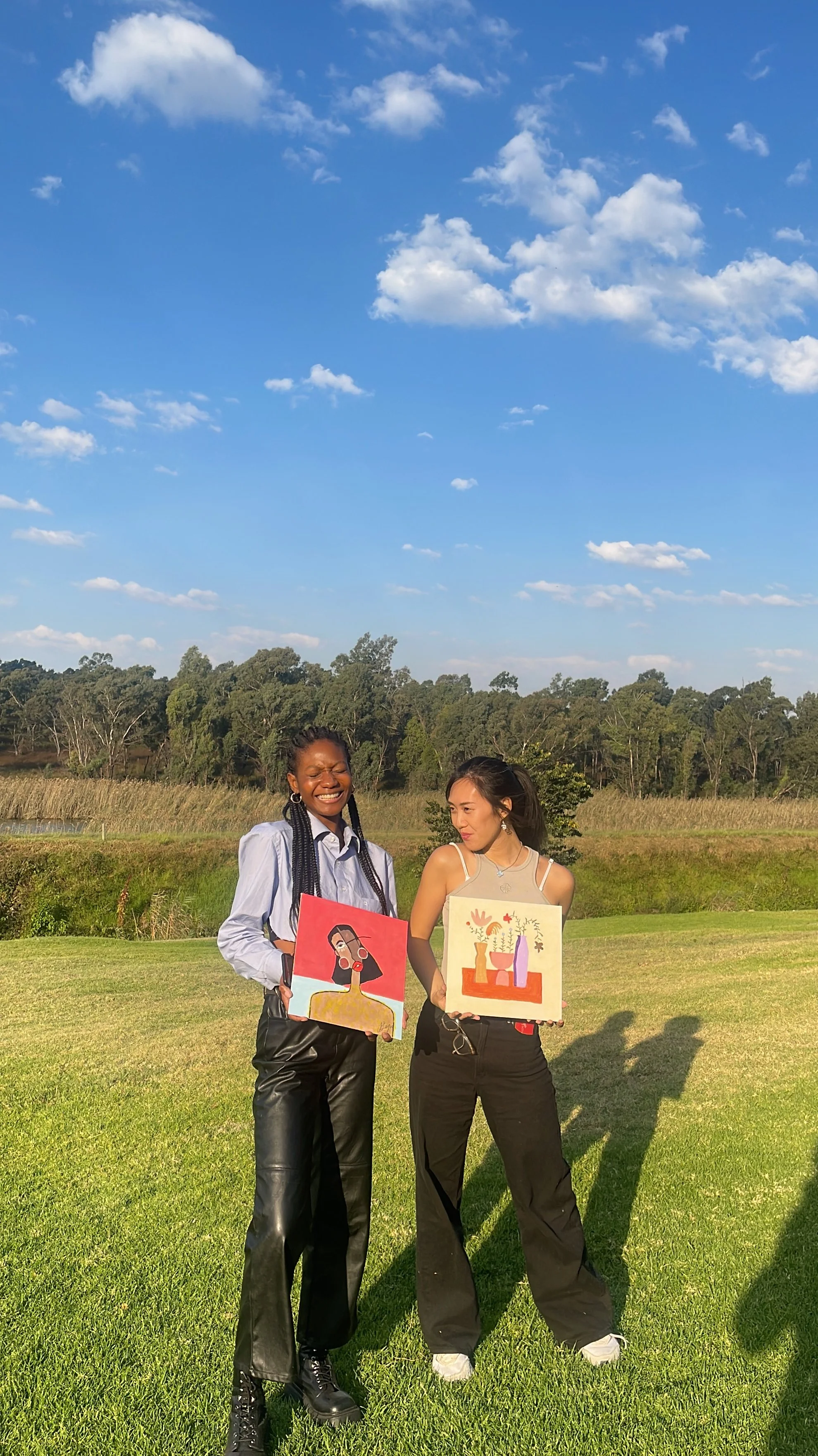 Two women standing outdoors on a grassy area, holding colorful art prints, smiling and looking at each other, with a backdrop of trees, open field, and a bright blue sky with scattered clouds.
