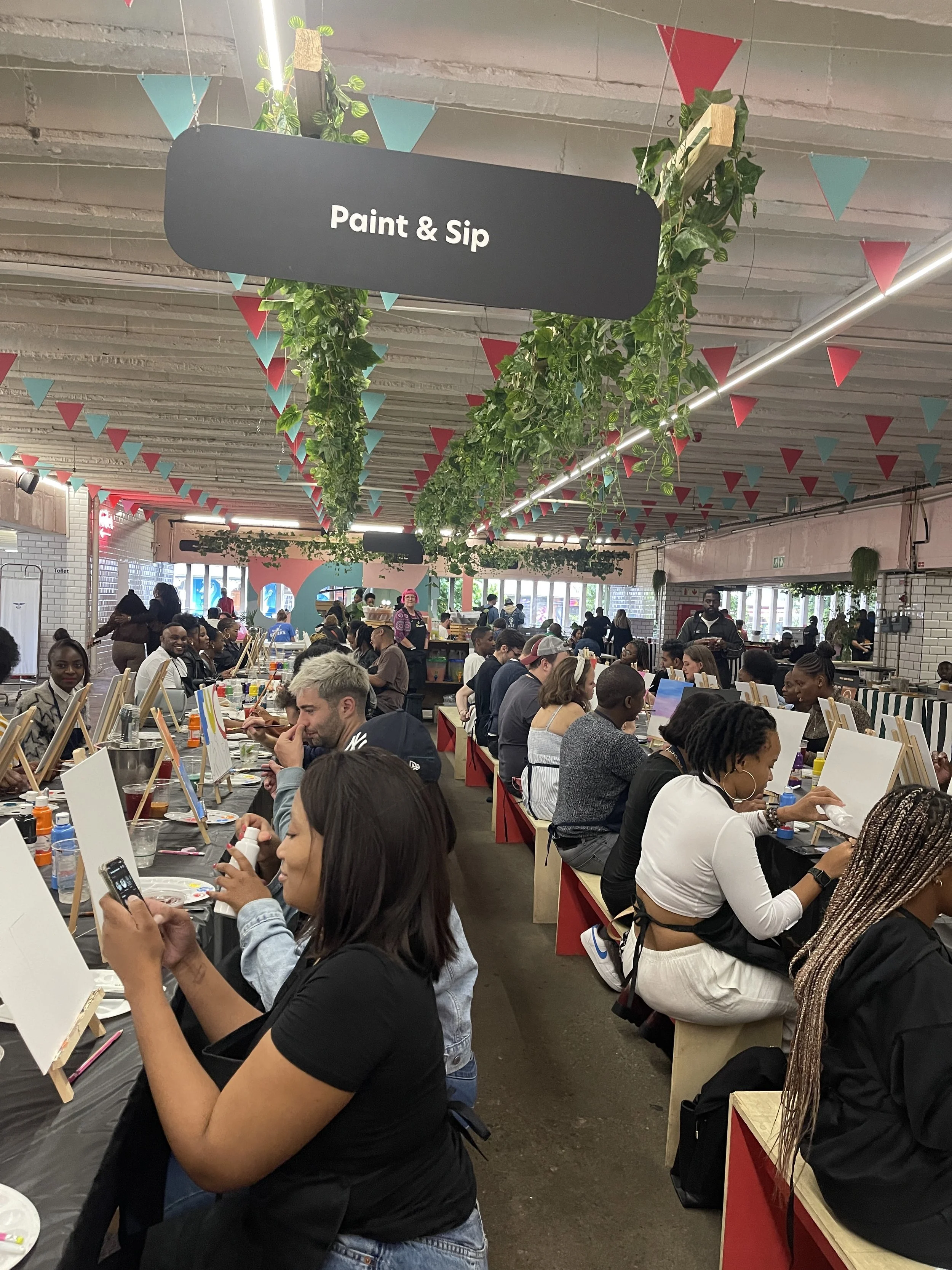 Group of people painting at long tables inside a decorated venue with bunting and hanging plants, sign reads 'Paint & Sip.'