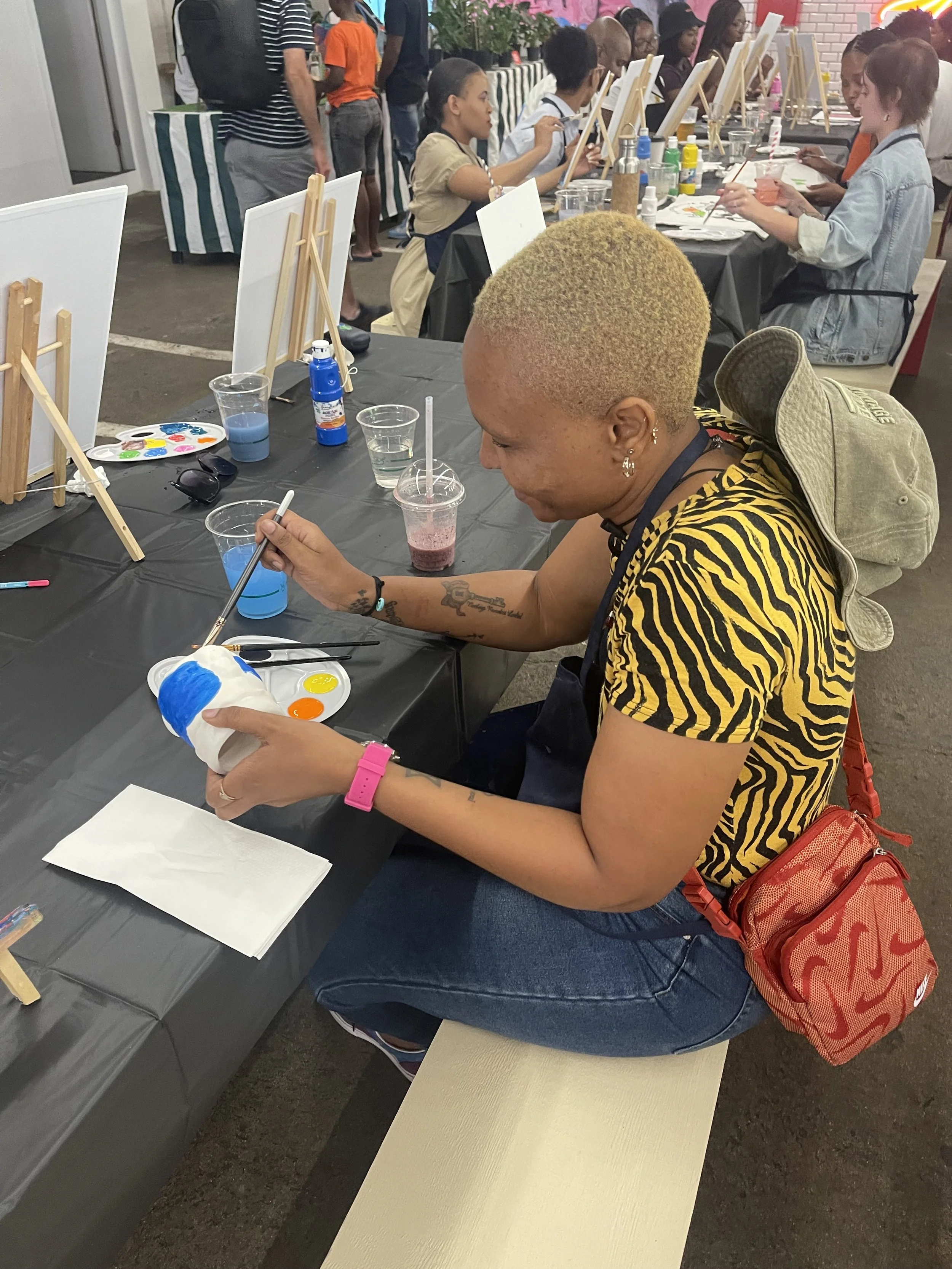 A group of people are seated at a long table participating in a painting activity, each with a canvas, paint, and brushes, while some stand in the background and observe.