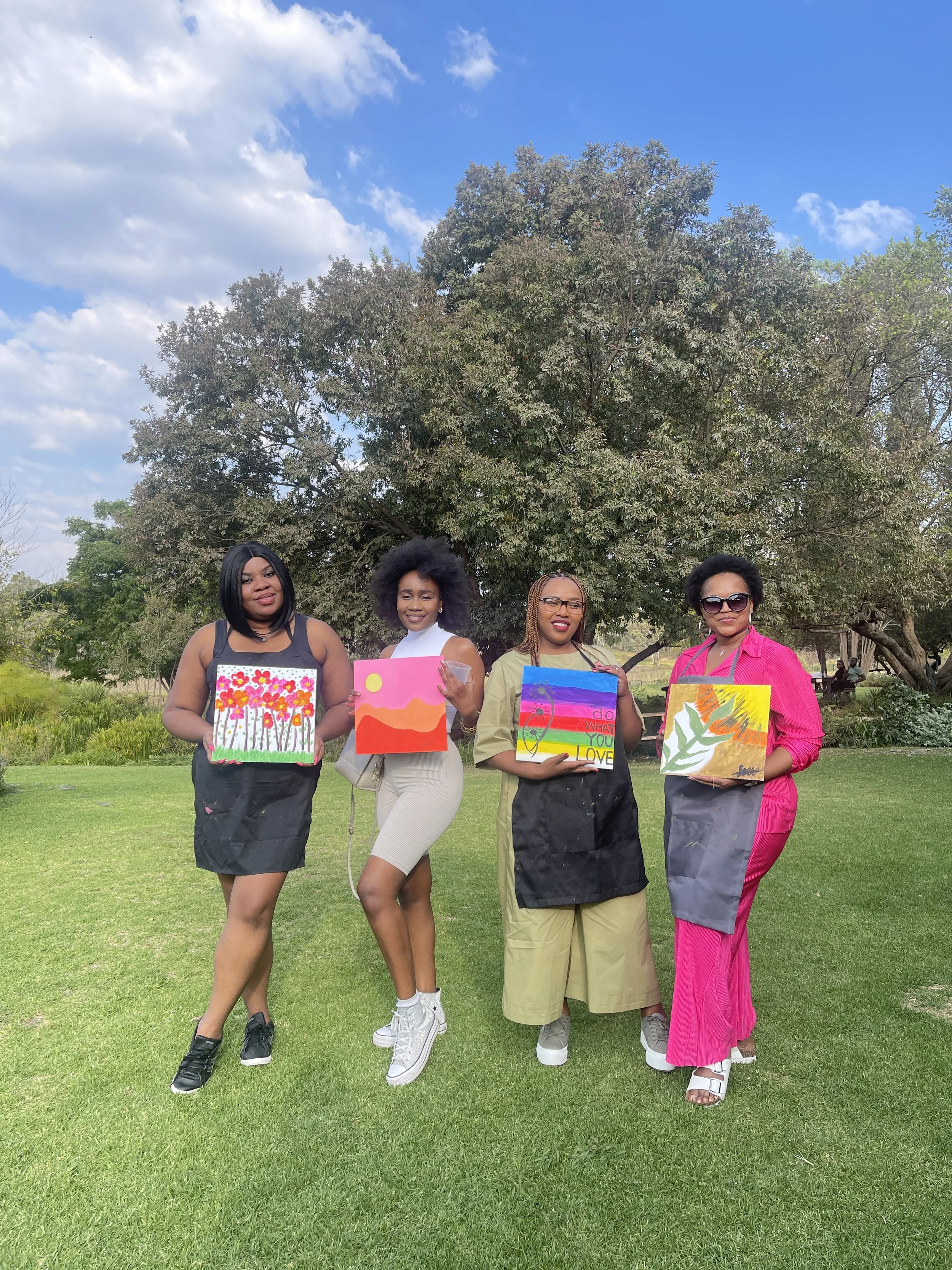 Four women standing outdoors on grass, each holding colorful paintings, with trees and a blue sky with clouds in the background.