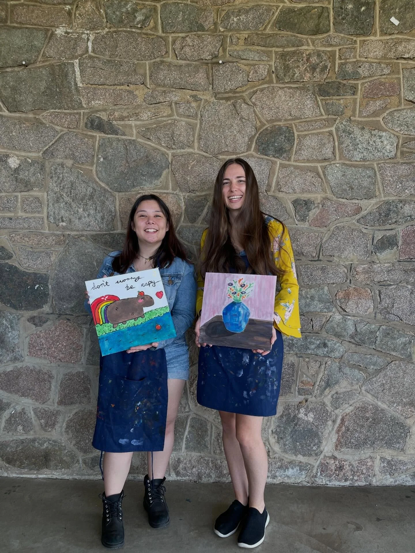 Two young women are standing in front of a stone wall, each holding a colorful painting. The woman on the left has dark hair and is wearing a denim jacket, shorts, and black boots. Her painting features a bear with a rainbow and the words 'don't worr