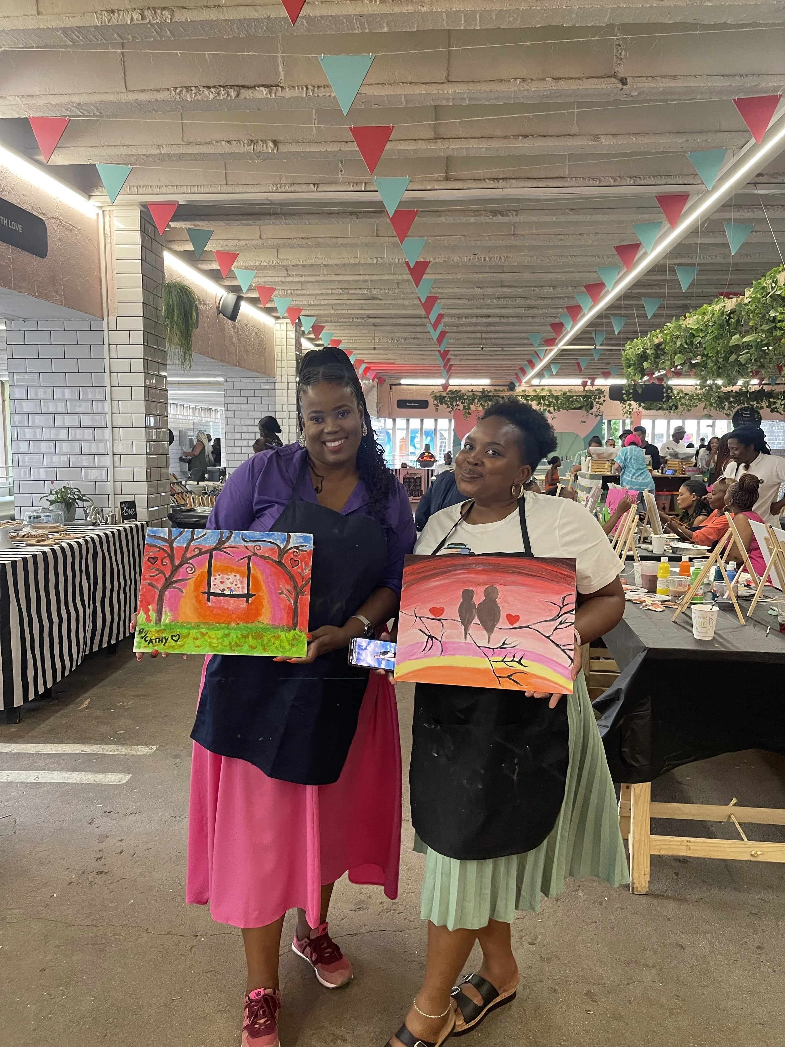 Two women standing inside a busy art market, holding colorful paintings of trees and a sunset with silhouettes of a couple and hearts. The woman on the left has dark hair in braids, wearing a purple shirt and a pink skirt. The woman on the right has 