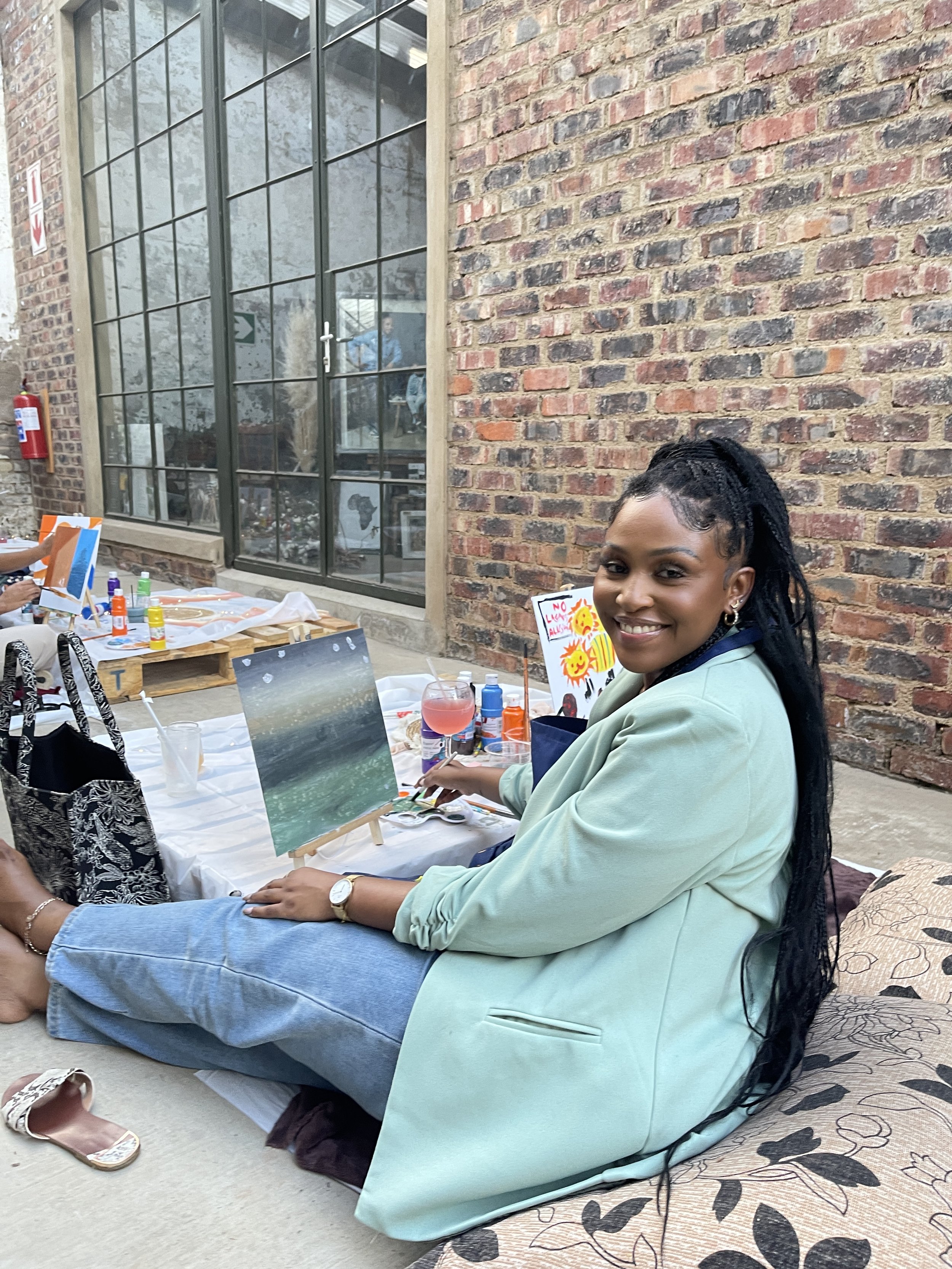 A woman with long black hair styled in twists, smiling, sitting cross-legged on the floor, painting on a small canvas set on an easel. She is wearing a light green blazer and blue jeans. In front of her on the table are bottles of paint, brushes, and