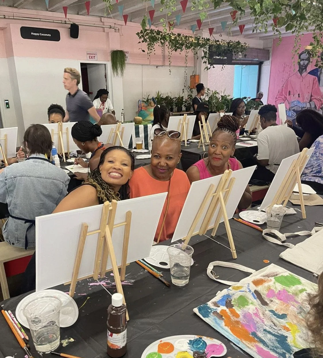A group of women participating in a painting class at a colorful studio with pink walls and art supplies. Three women are smiling at the front, sitting behind small easels. Several others are working on their paintings in the background, some wearing