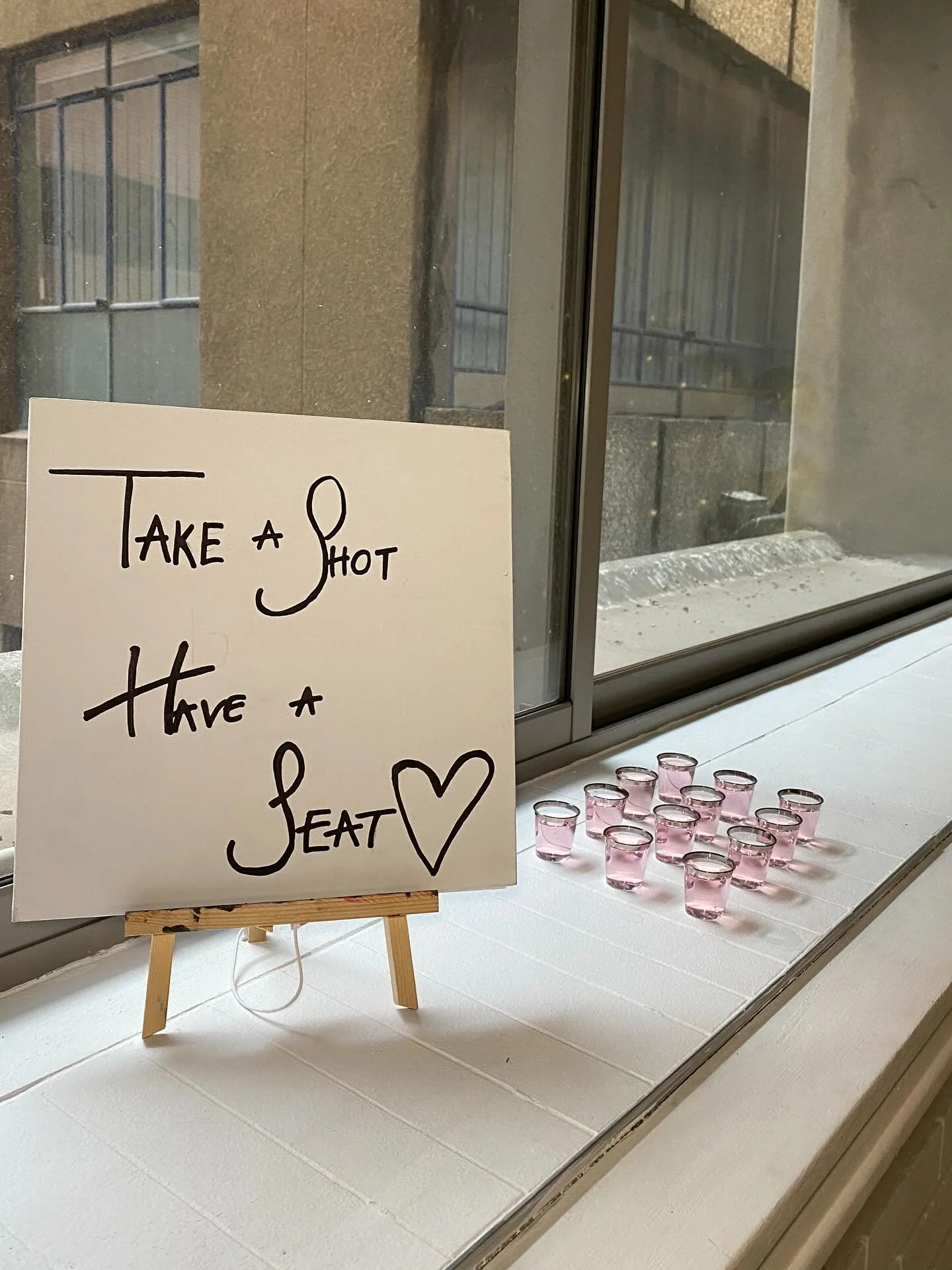 A white sign on a small wooden easel that says "Take a shot, have a seat" with a heart drawing. Next to the sign, there are small pink shot glasses filled with a drink, arranged in rows on a white surface, inside a room with a large window showing an