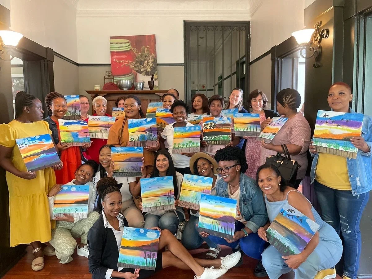 Group of women and girls indoors, holding colorful landscape paintings they created, smiling for the camera.