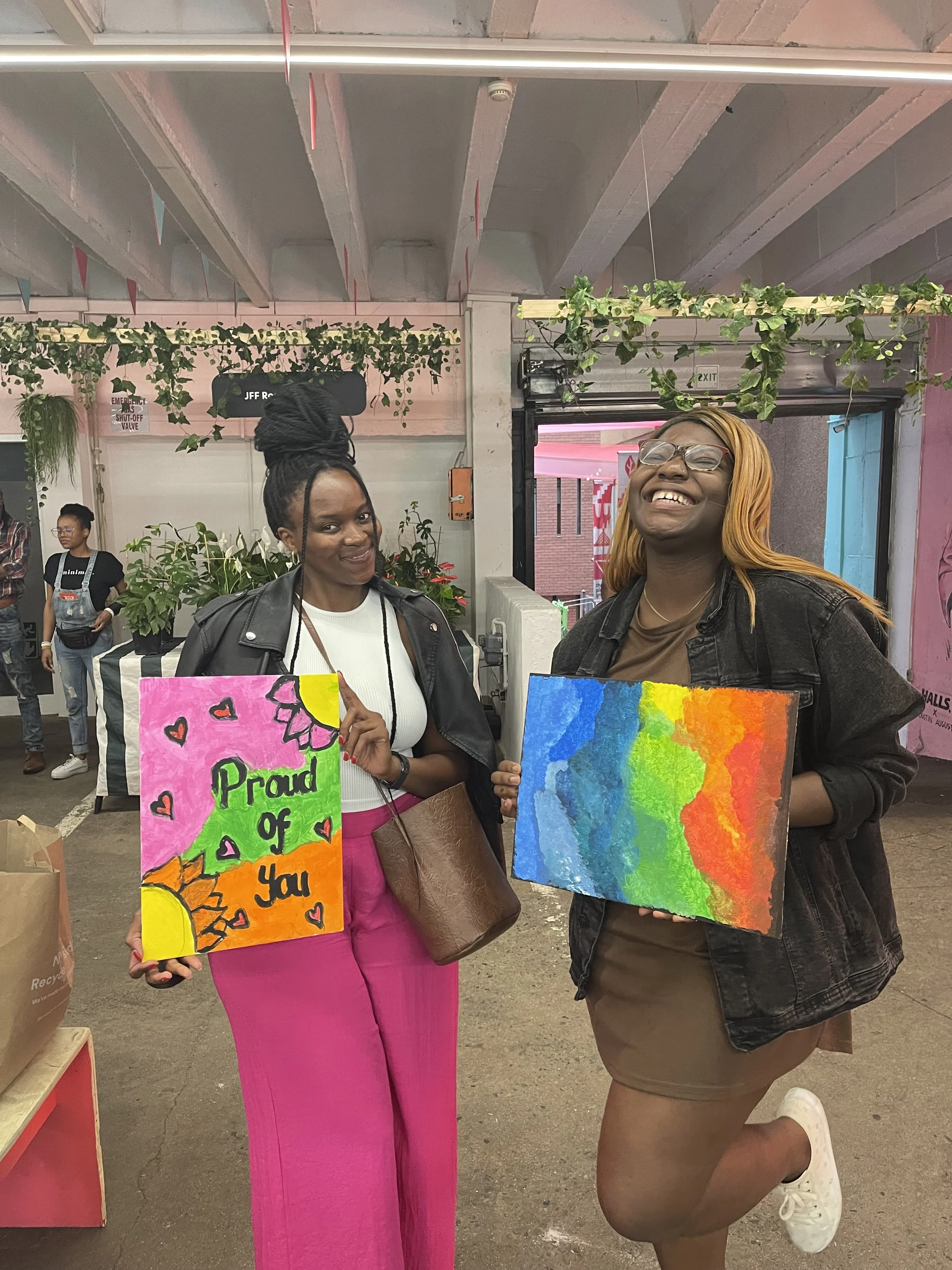 Two women standing inside a building, holding colorful signs. One woman is smiling and wearing glasses, with blonde hair, dressed in a brown top and black jacket. The other woman has darker hair styled in braids with a top knot, wearing a white top a