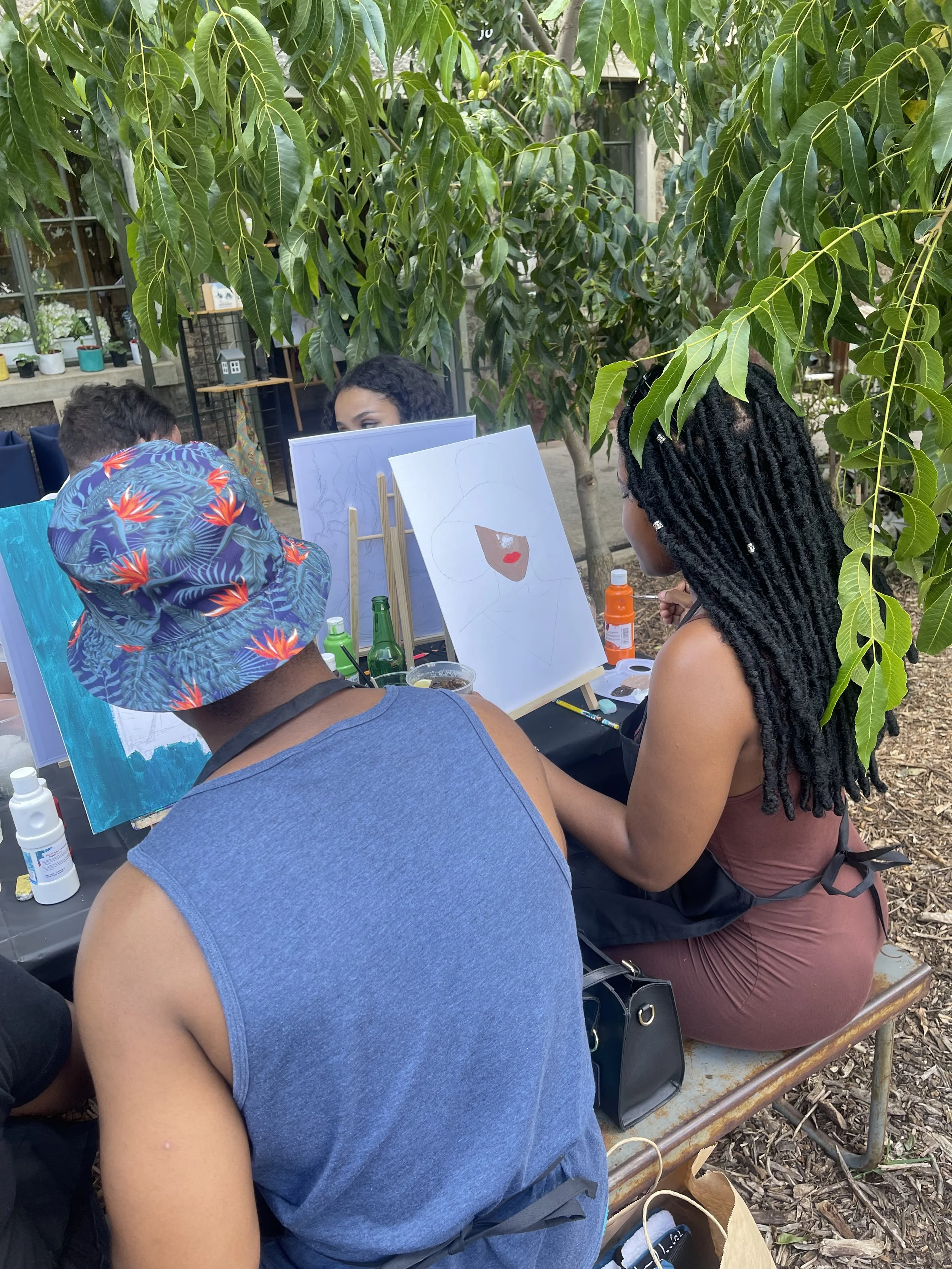 People painting portraits outdoors under trees, with art supplies and paintings on display.