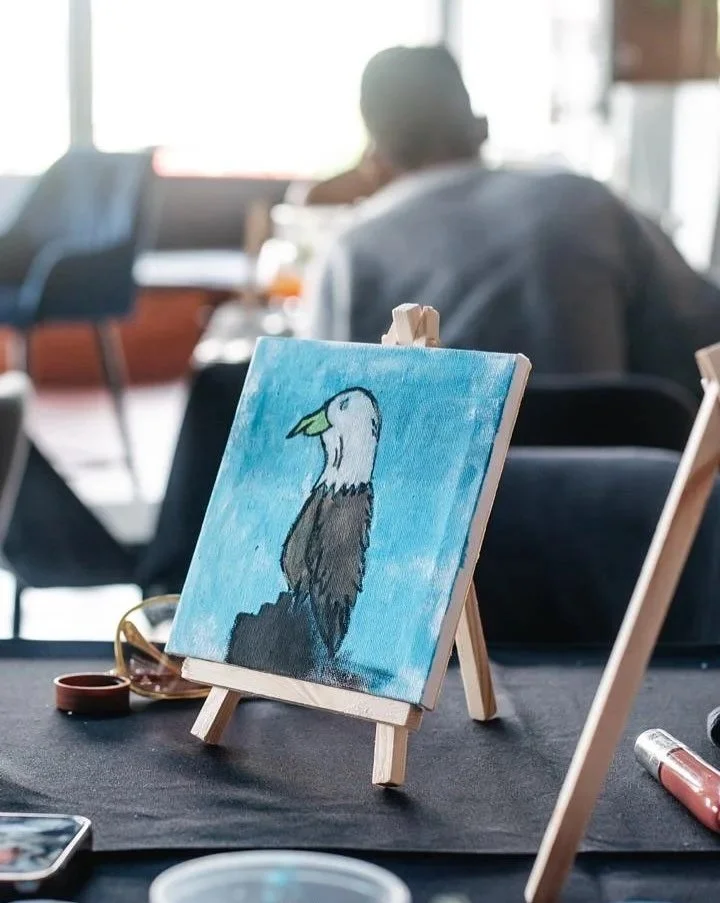 A small canvas painting of a bird with a white head and dark body on a mini easel, set on a table with art supplies, with a blurred person in the background.