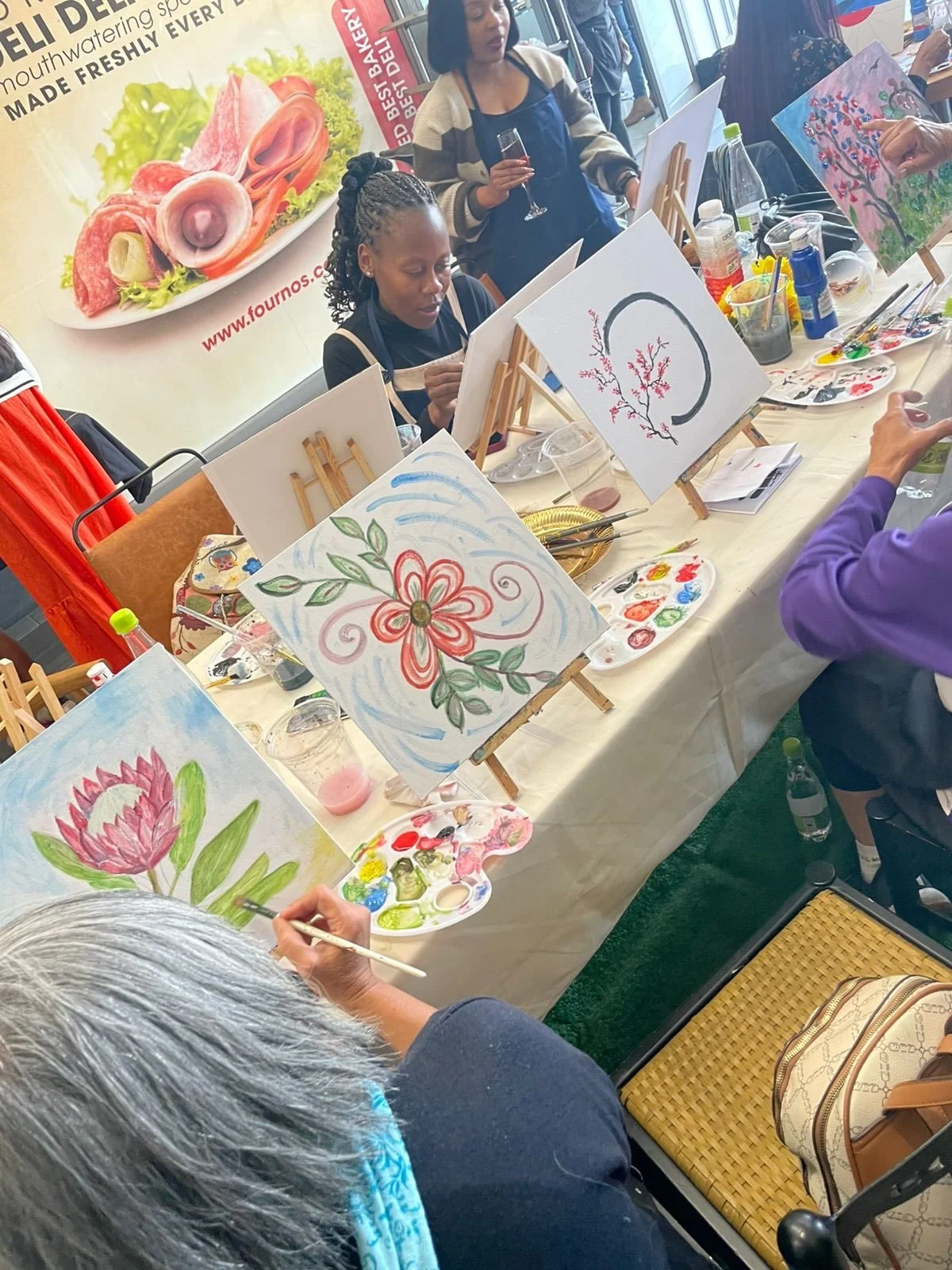 People participating in a painting class, creating floral artwork on canvases, with painting supplies and refreshments on the table.