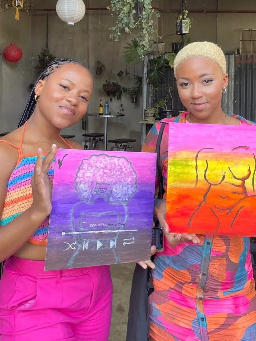 Two women holding colorful paintings, smiling indoors, with plants, hanging lanterns, and a metal wall in the background.