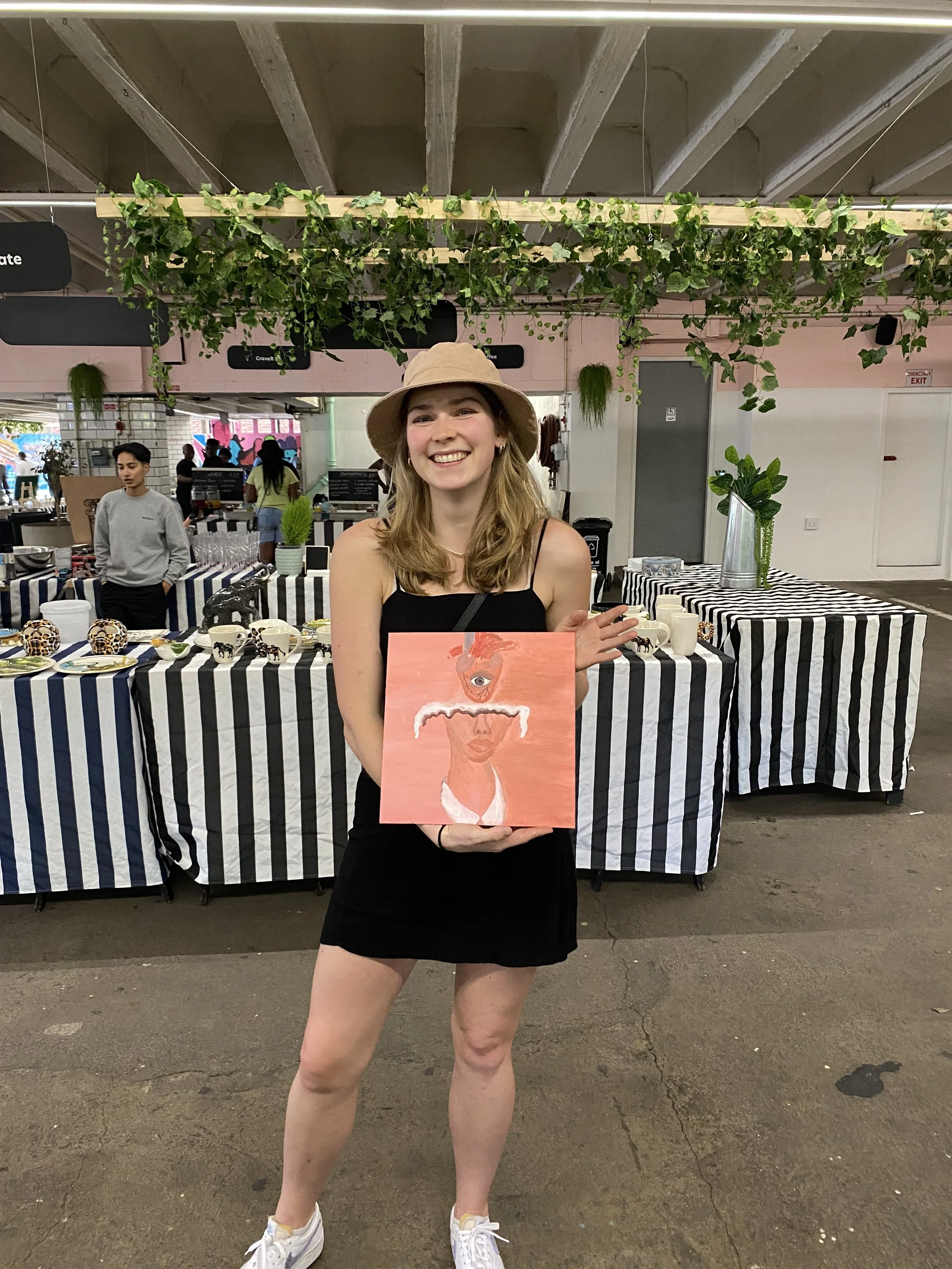 A young woman with long, wavy blonde hair, wearing a beige hat, black tank top, and black shorts, is standing indoors holding a painting of a face with an eye on a peach-colored background. The background shows a market or event space with tables cov