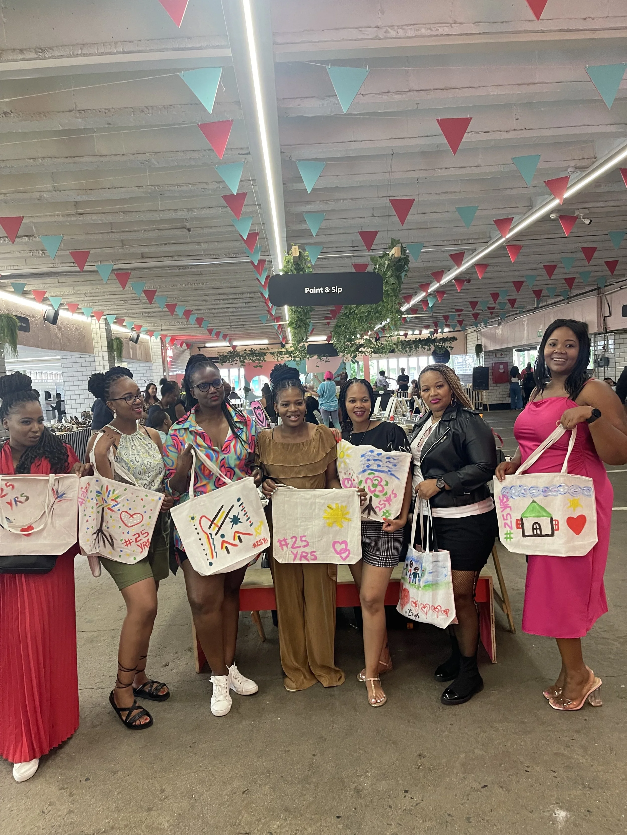 A group of women standing indoors, holding colorful tote bags with various drawings and messages, in celebration of a 25th anniversary.