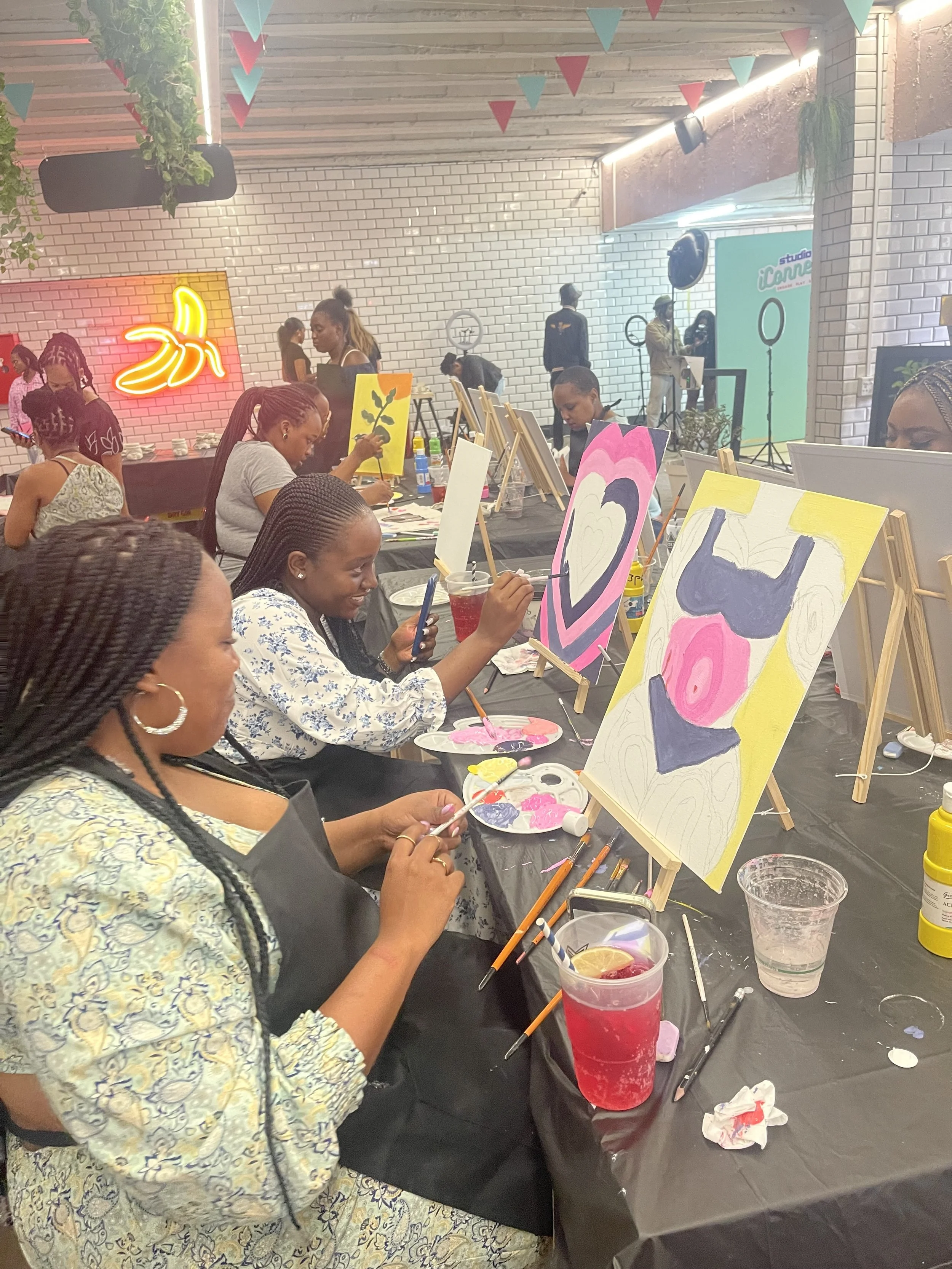 A group of women painting colorful abstract art on canvases at an indoor art party. Some women are focused on their paintings, while others are chatting or looking at their phones. There are drinks, art supplies, and vibrant decorations, including a 