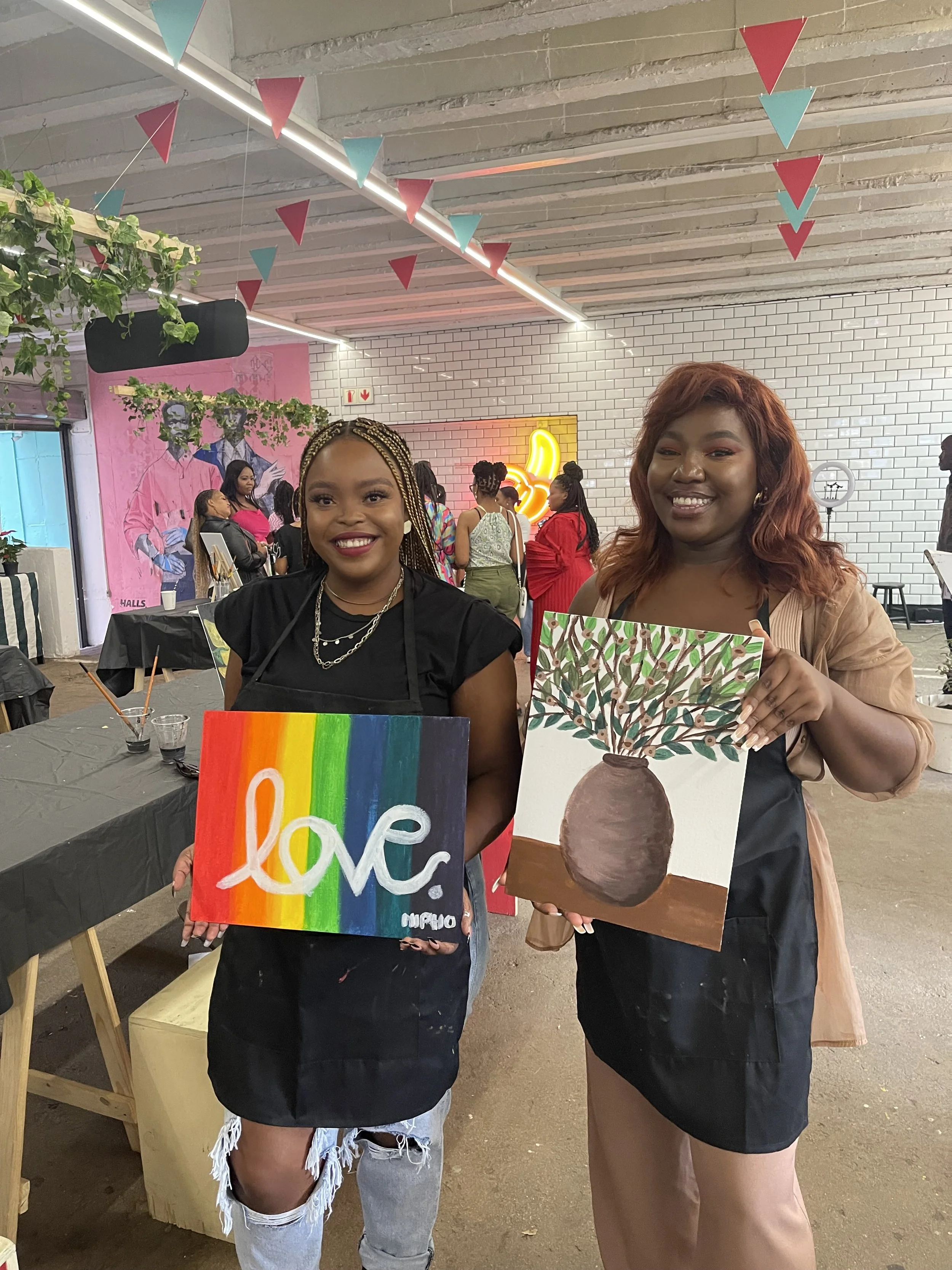 Two women smiling and holding paintings, surrounded by a lively, decorated indoor space with people in the background.