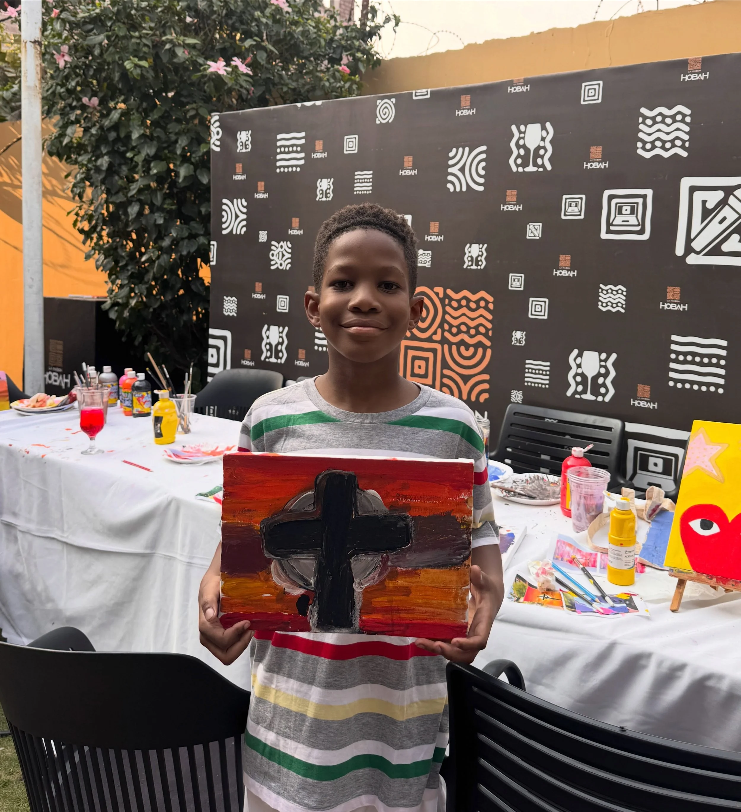 A young boy wearing a gray, green, yellow, and white striped shirt presents a colorful abstract painting on a canvas at an outdoor art event. Behind him is a large black backdrop with white and orange patterns, and a table with art supplies and drink