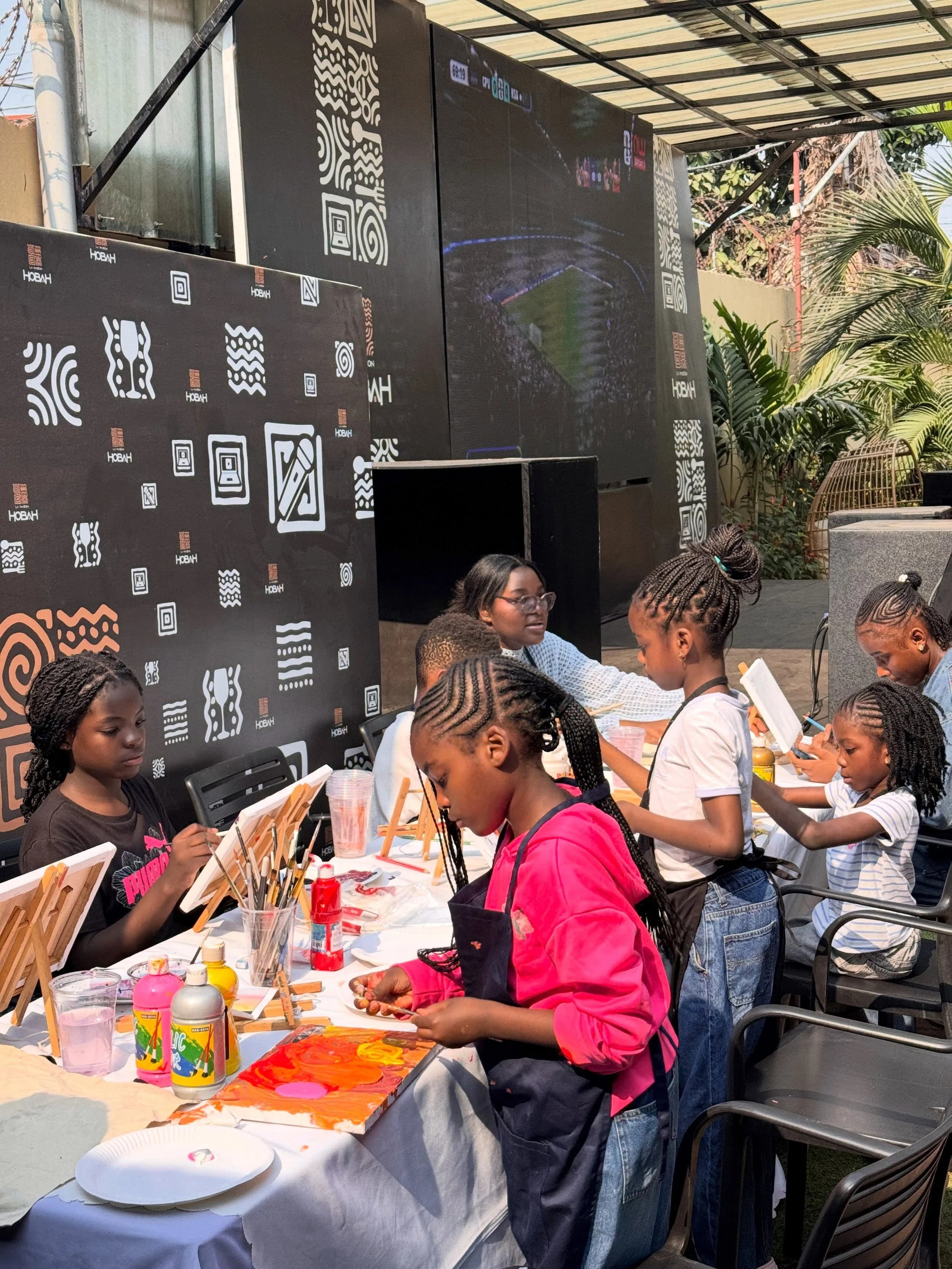 Children and adults painting and crafting at an outdoor art event, with art supplies such as paint bottles, brushes, and easels on the table, and a decorative black backdrop with white patterns.