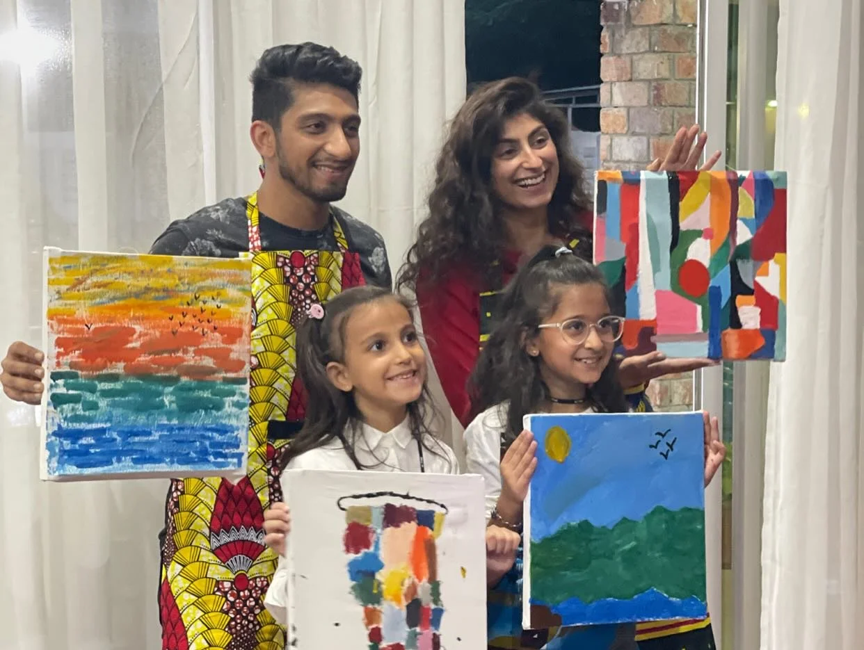 Group of five people, two adults and three children, holding colorful paintings in an indoor setting.