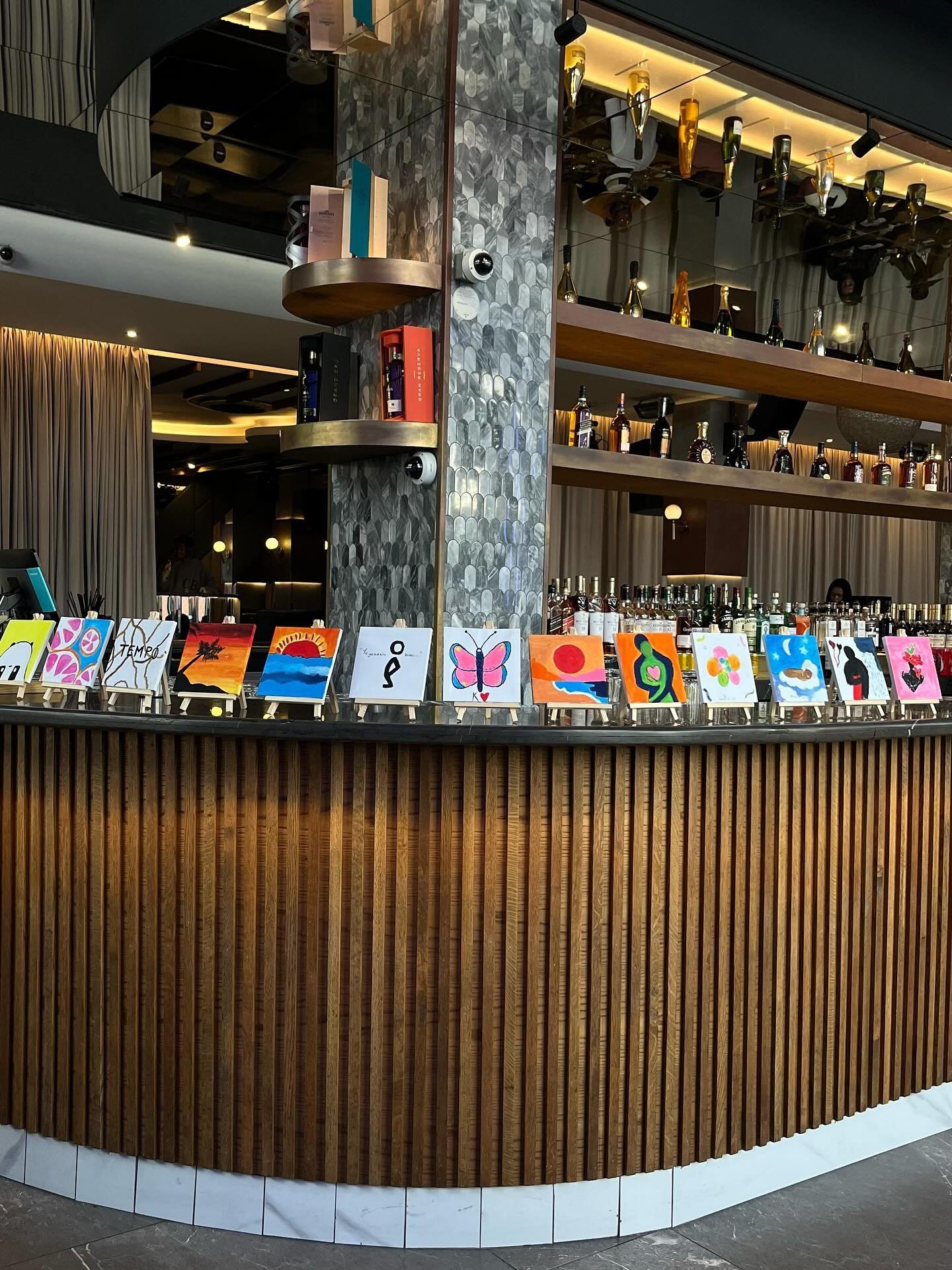 A bar with a wooden curved counter and art displays on small easels in front. Behind the bar, shelves hold bottles of alcohol, and above are decorative lights and bottles on a higher shelf. Curtain drapes are visible in the background.