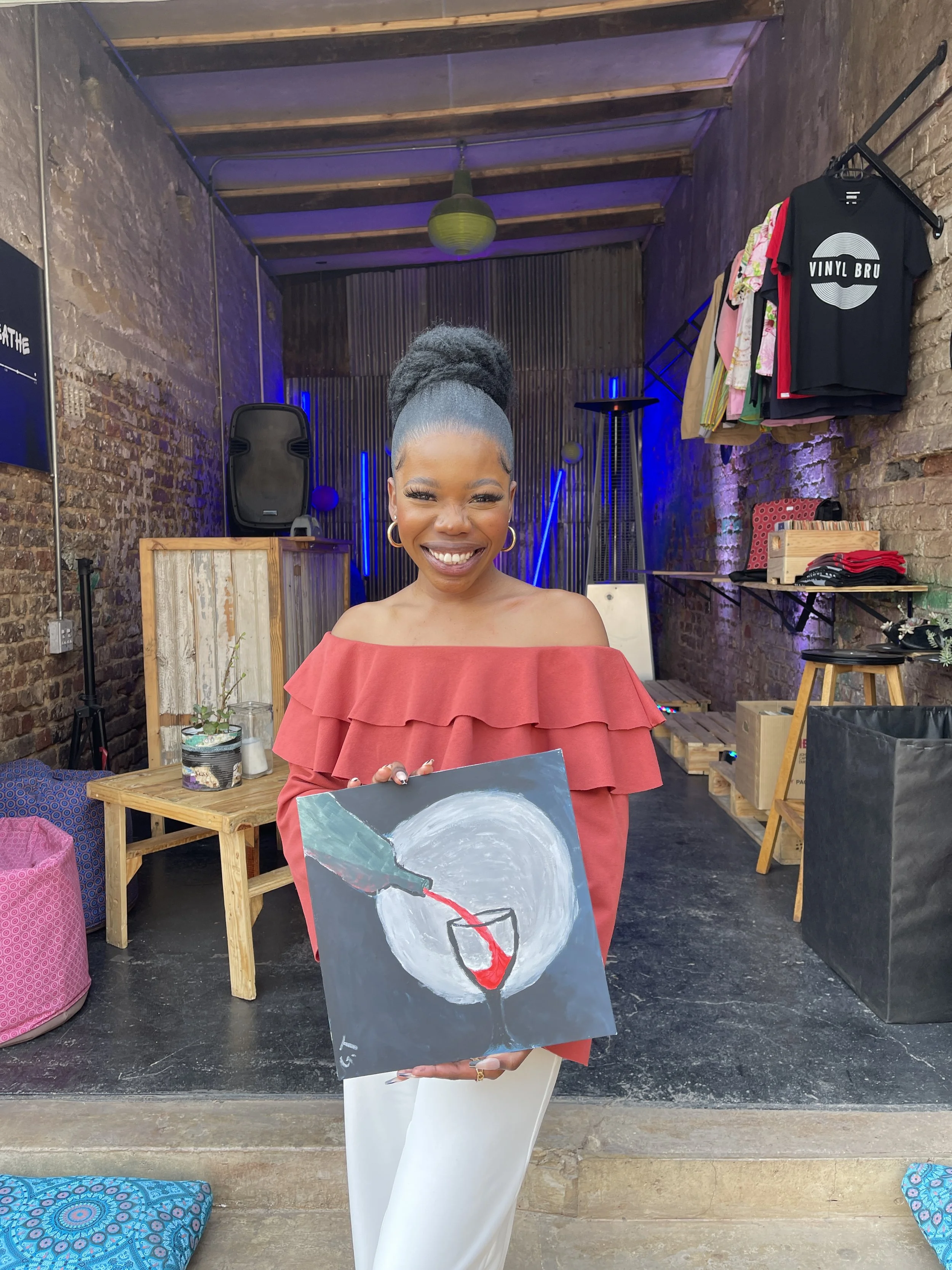 A woman smiling and holding a painting of a wine glass with red wine, standing in an art studio or gallery with exposed brick walls and artwork.