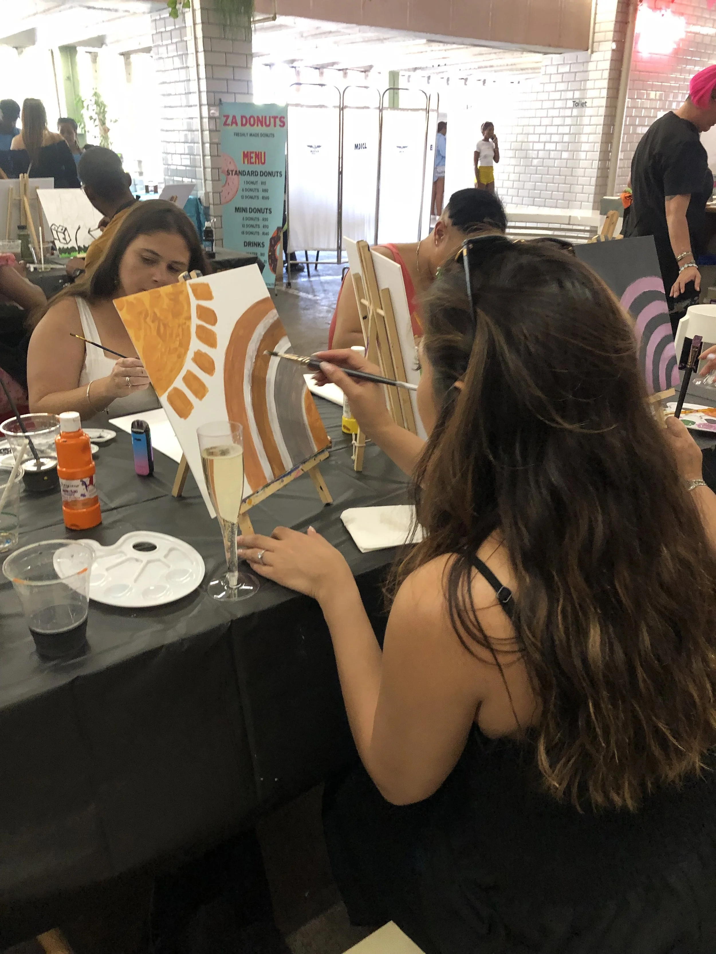 People painting canvases at an art event in an indoor space with white brick walls and a black table.