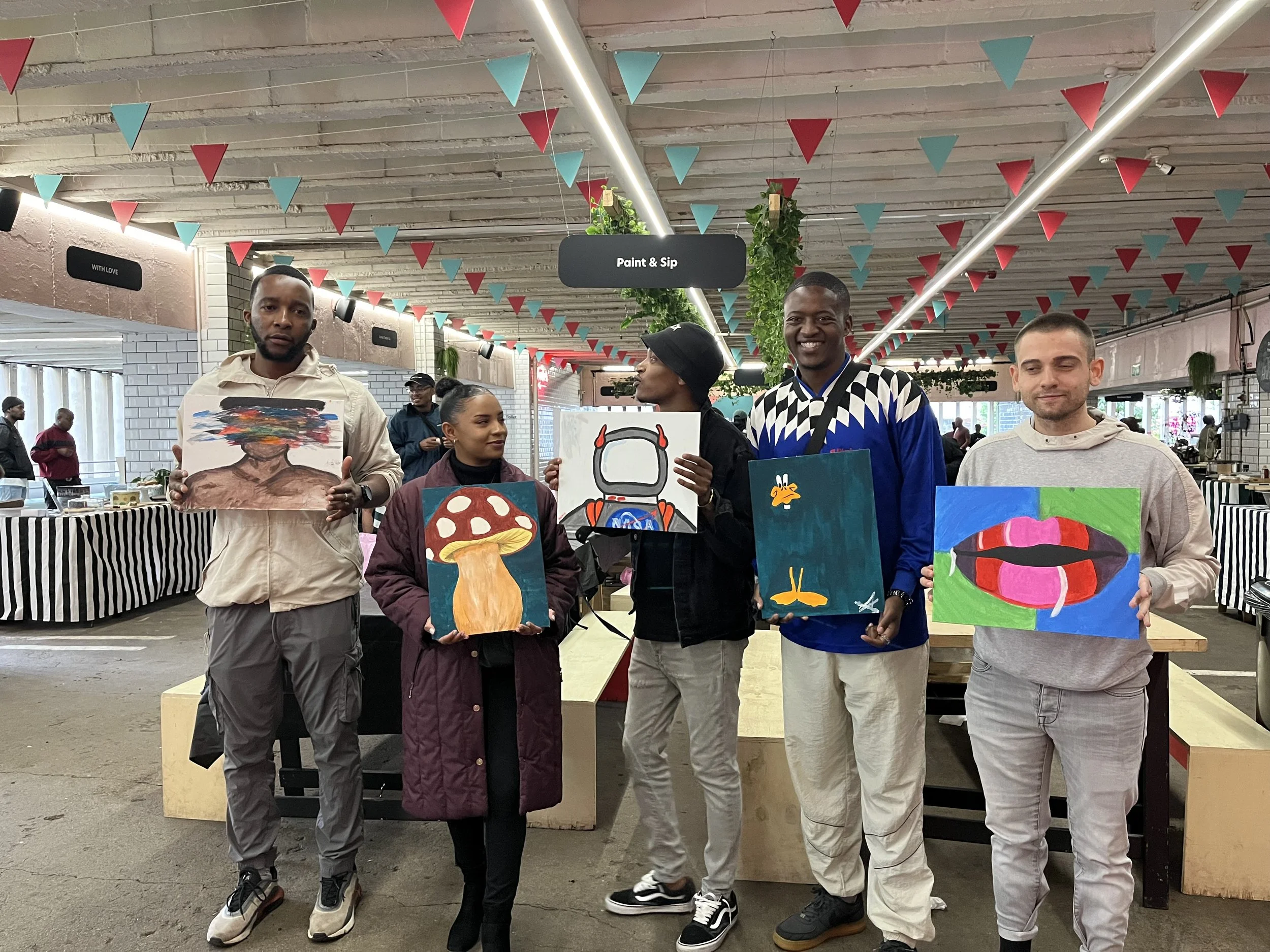 Five people standing indoors, each holding a colorful painting. The ceiling is decorated with red and blue triangular pennants. There are plants hanging from the ceiling and a black sign reading "Paint & Sip" hangs above them. In the background, othe