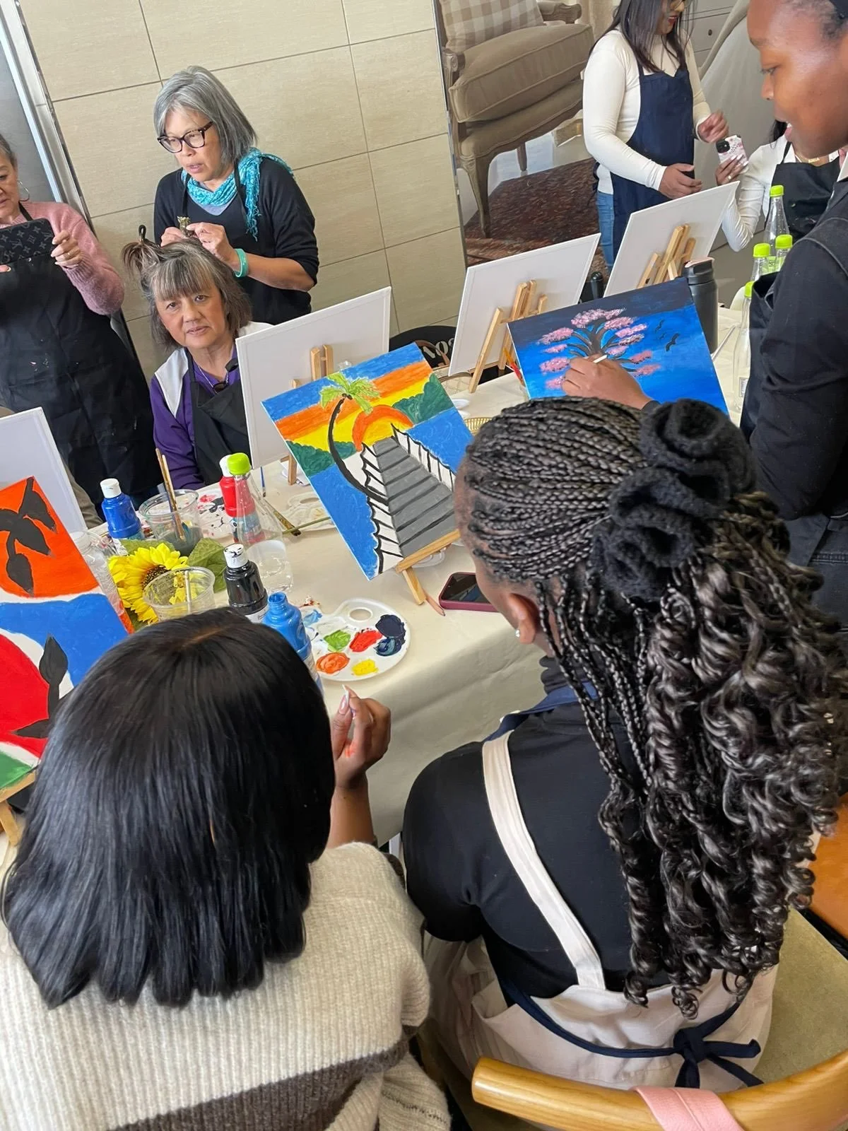 People participating in a paint and sip event, creating colorful paintings of sunset views, cherry blossom trees, and abstract designs on canvas. The table has art supplies, drinks, and flowers.