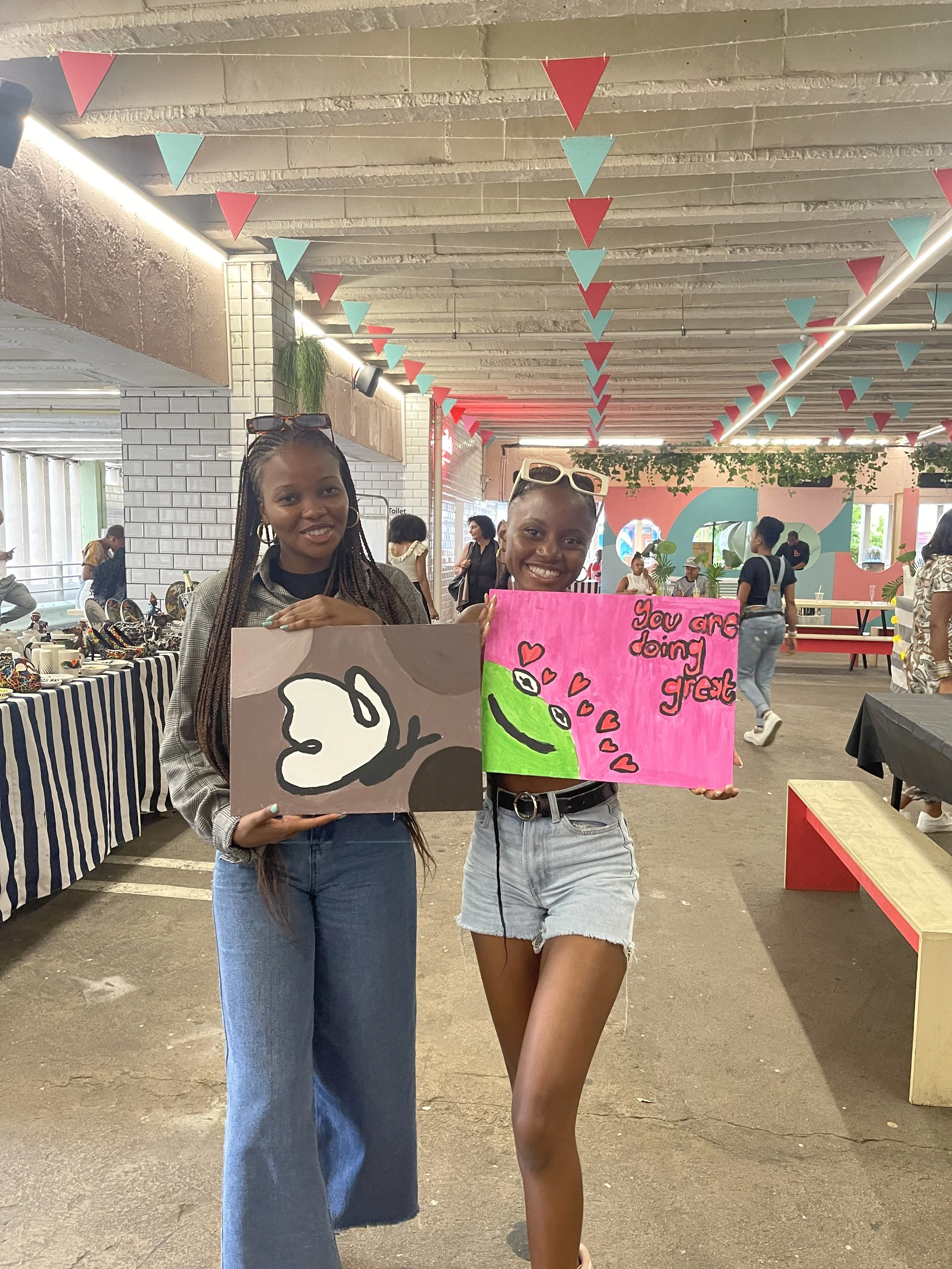 Two young women at a marketplace or event holding handmade signs with positive messages, one with artwork of a butterfly and the other with a frog surrounded by hearts, smiling at the camera.