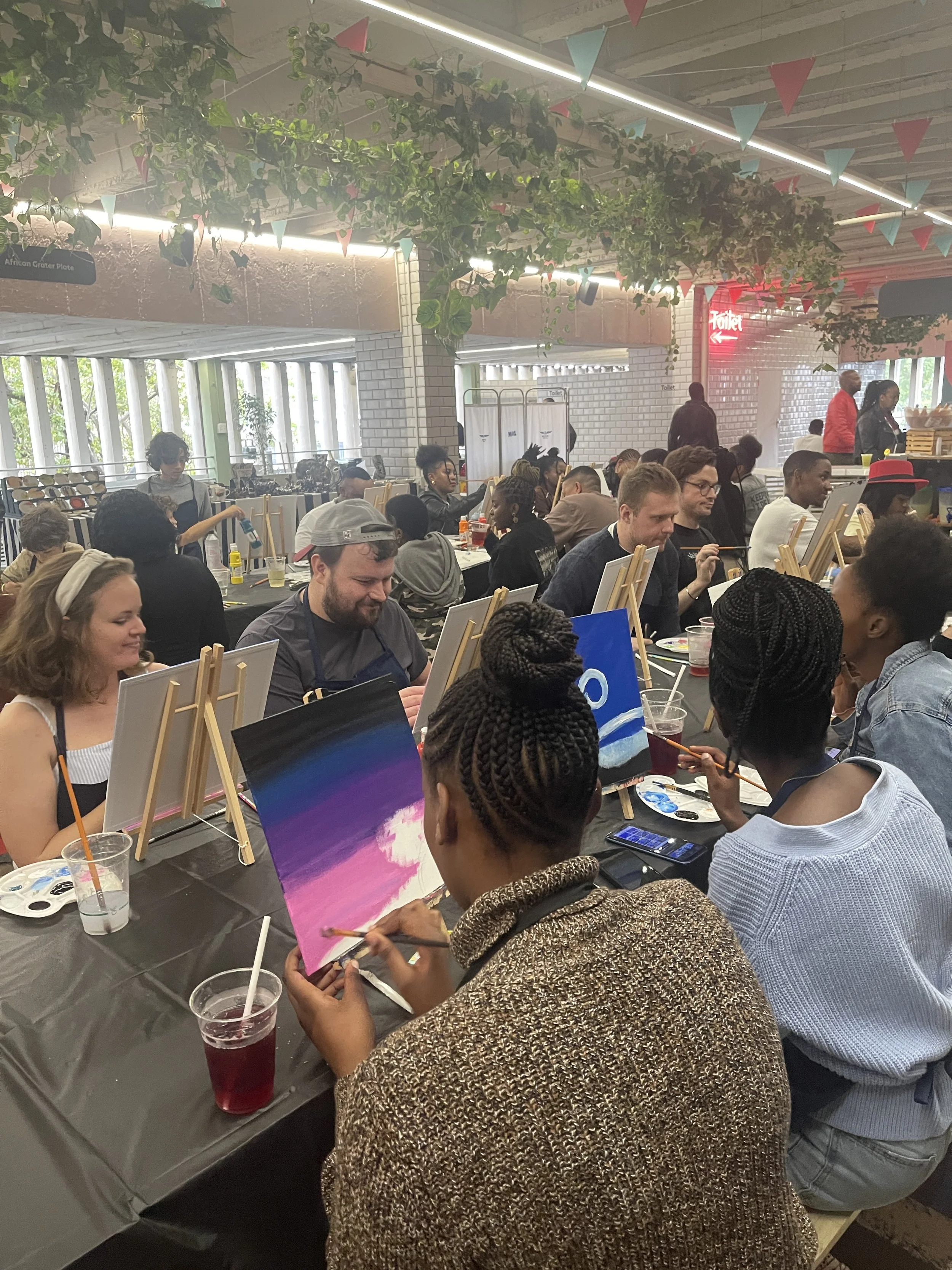 People sitting at a long table painting on small canvases. The table is covered with a black cloth and drinks are visible. The room is decorated with hanging greenery and triangular flags. Some individuals are focused on their art, while others are e