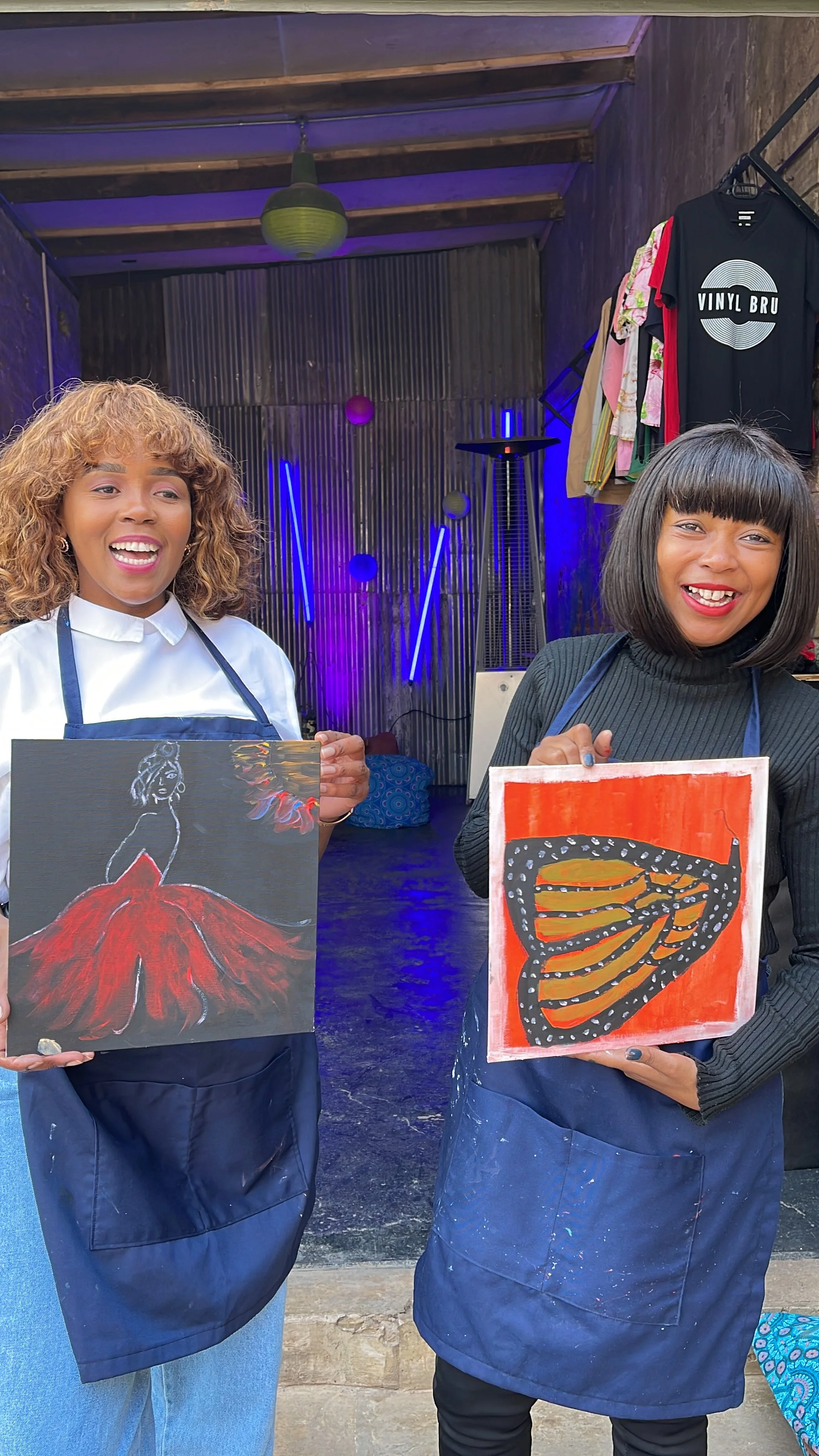 Two women wearing aprons holding paintings, smiling indoors with clothing hanging on the wall and neon lights in the background.