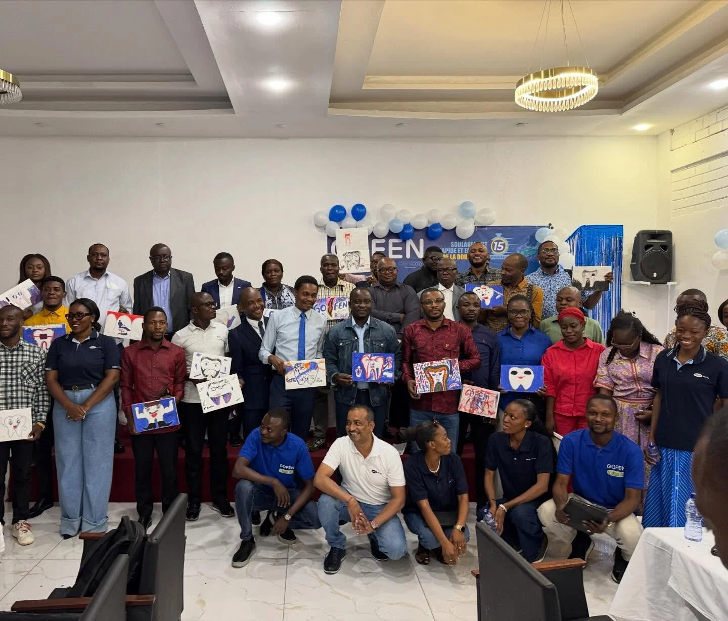 Group photo of people at an event, some holding colorful drawings of smiles, teeth, and dental health themes, with a backdrop decorated with blue and white balloons and a banner celebrating 15 years.