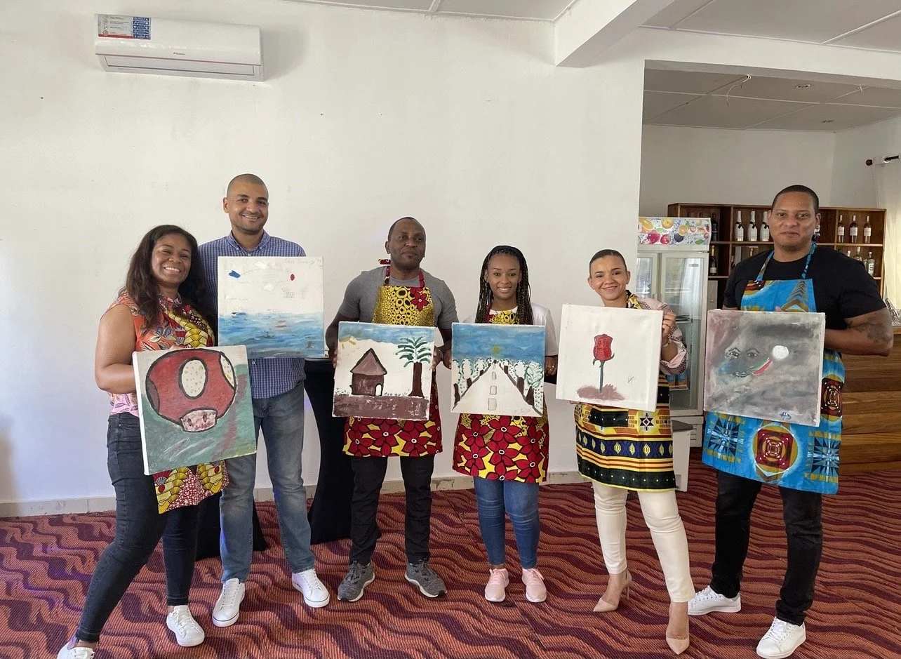 Group of six people holding paintings during a painting class, standing on a red patterned carpet in a room with white walls and an air conditioner.
