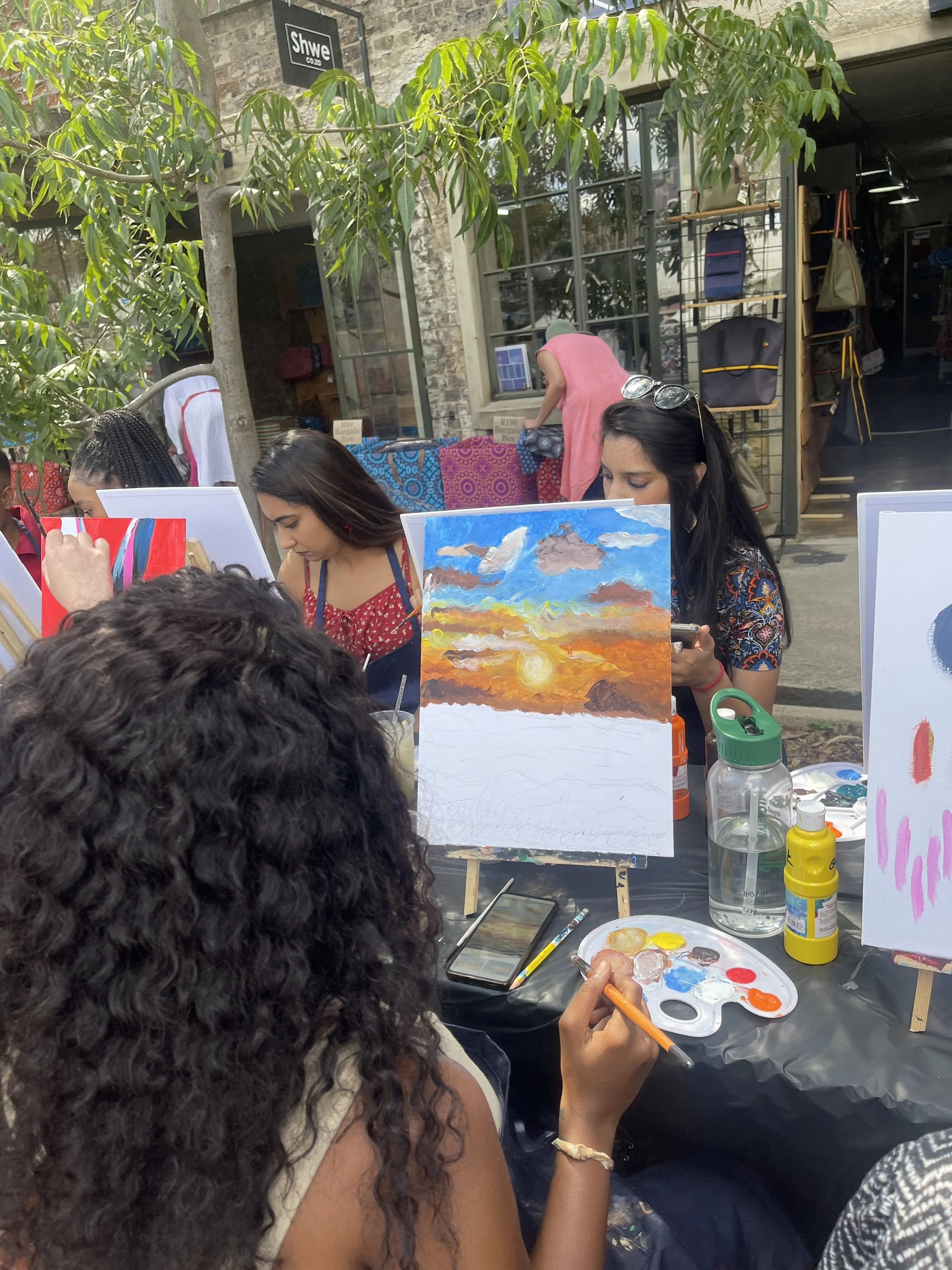 People painting outside with art supplies on tables, including a colorful sunset painting, in front of a building with large windows and a sign that says 'SHWE'.