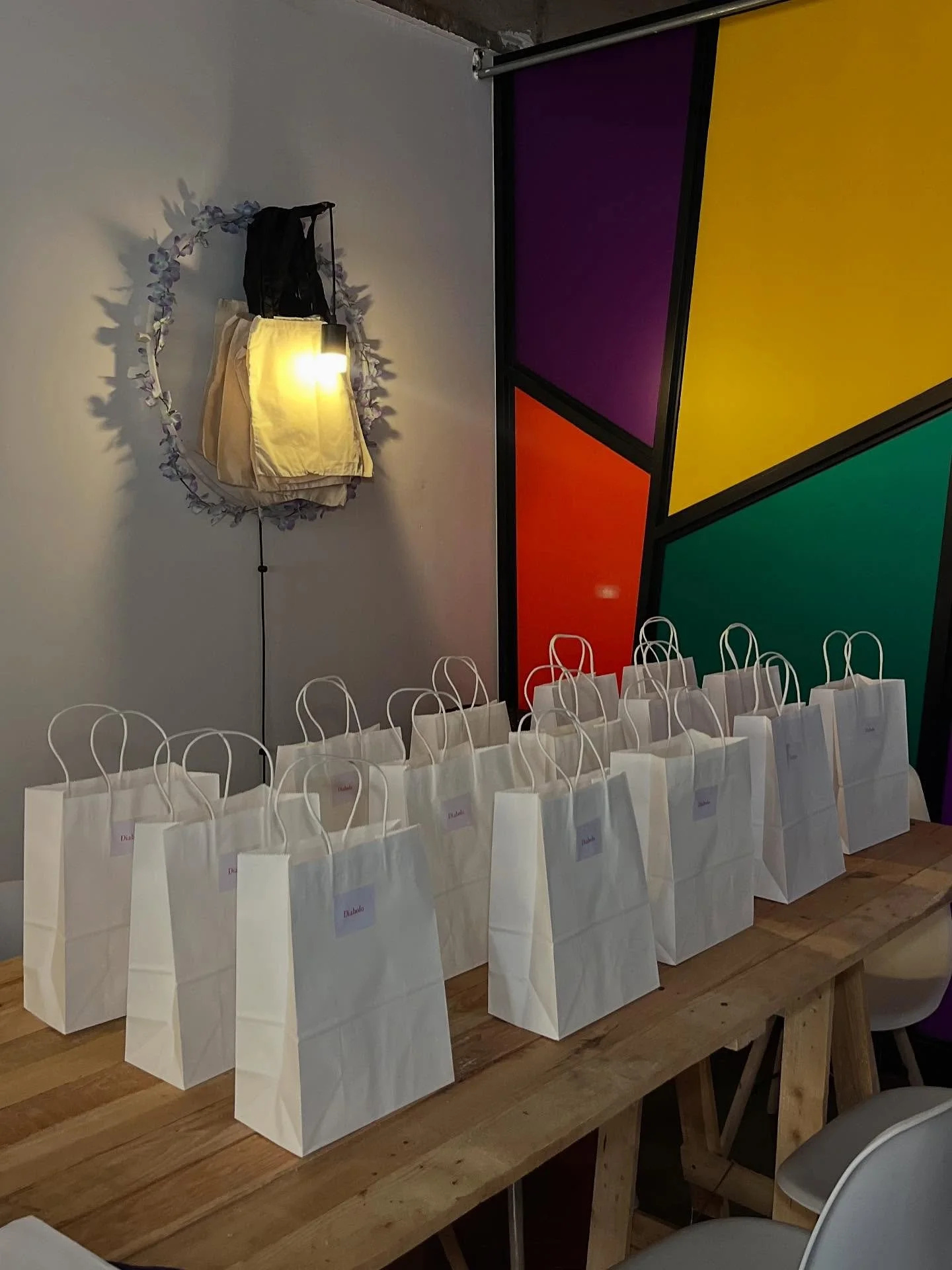 White gift bags with handles lined up on a wooden table, each with a label reading "Diatta". In the background, a wall decorated with a floral wreath and a yellow, purple, red, and green panel.