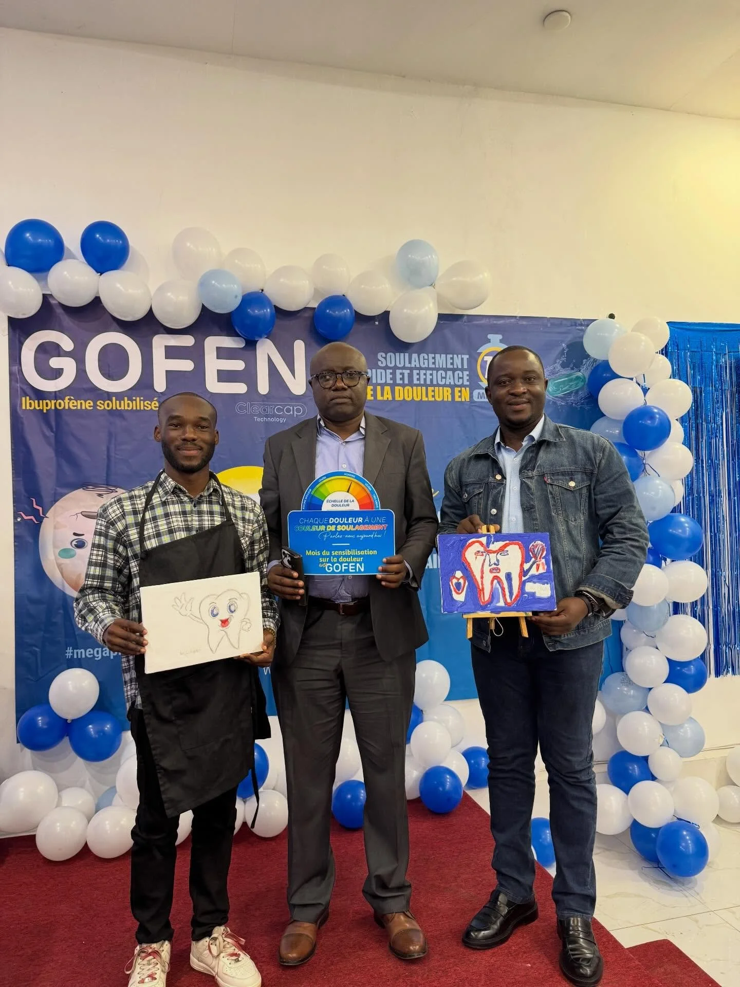 Three men posing in front of a blue and white balloon backdrop at a health awareness event. The man on the left is holding a drawing of a smiling tooth. The man in the middle is holding a sign with a colorful tooth illustration. The man on the right 