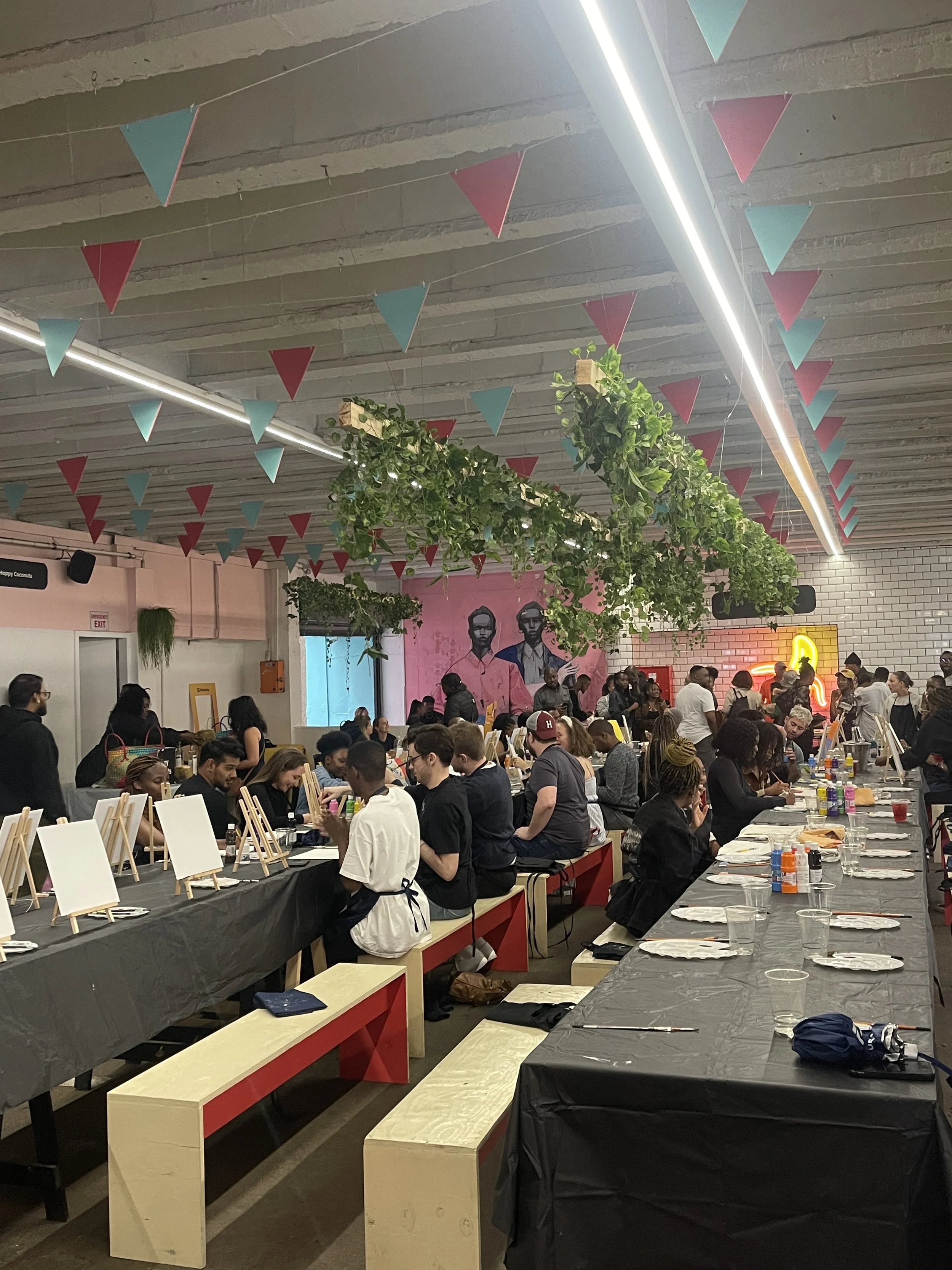 People gathered inside a decorated event space, preparing for a painting activity, with canvases on tables, leafy hanging plants, and colorful triangular banners hanging from the ceiling.