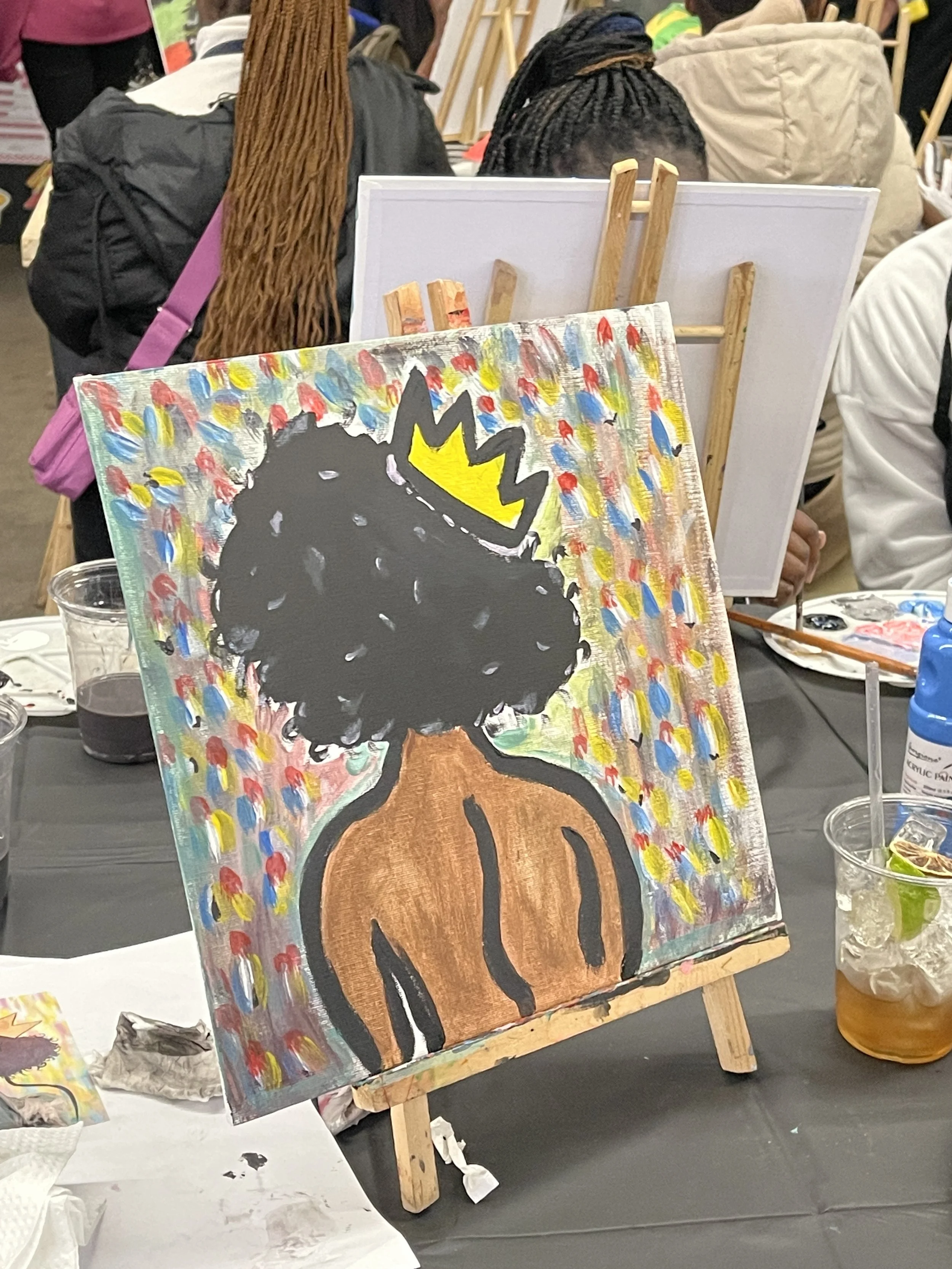 Painting of a person with black, curly hair wearing a brown top, with colorful abstract background on an easel at an art event.