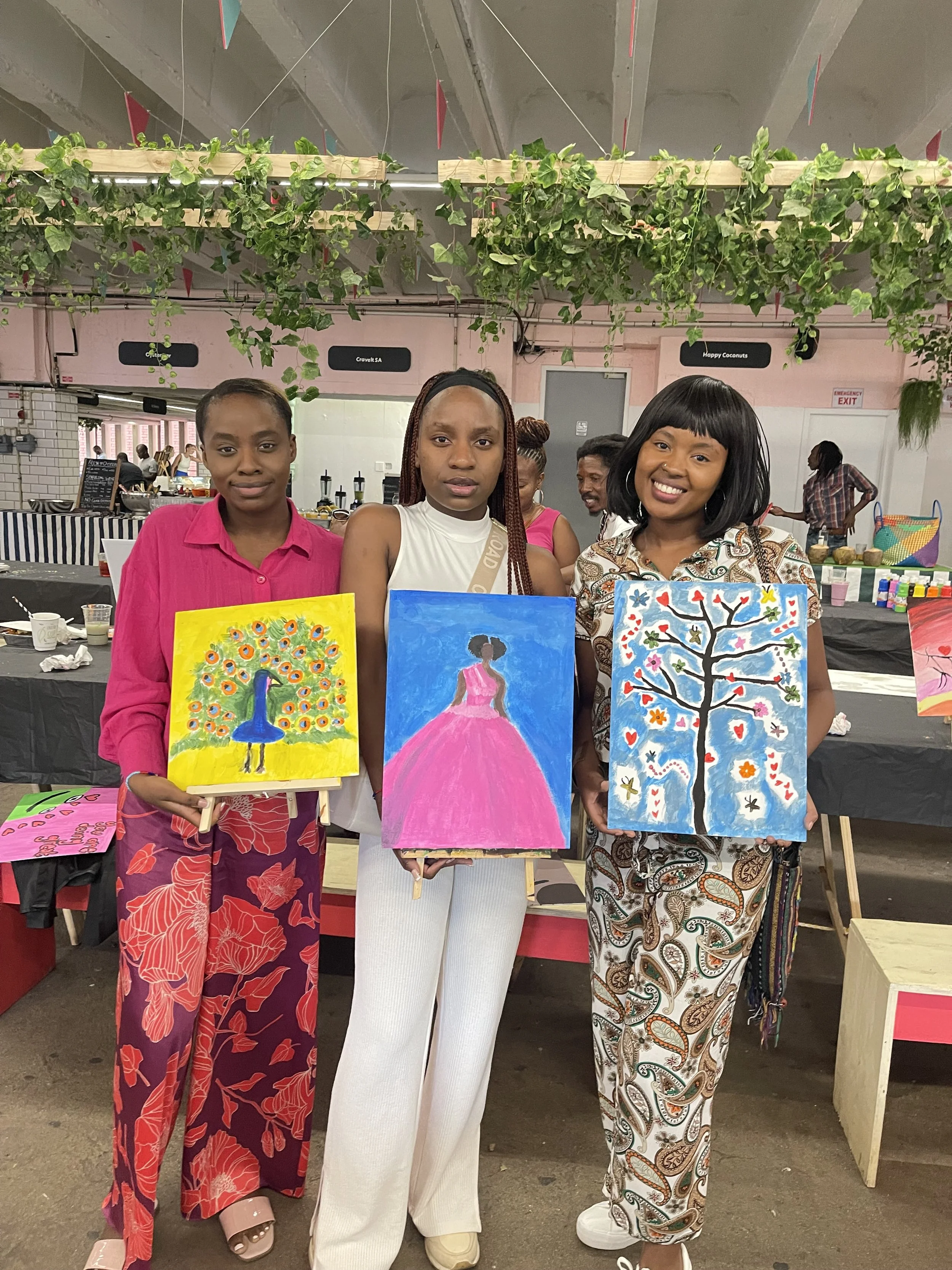 Three women standing side by side indoors, each holding a painting on a small easel, with a man and woman in the background and various items on tables.