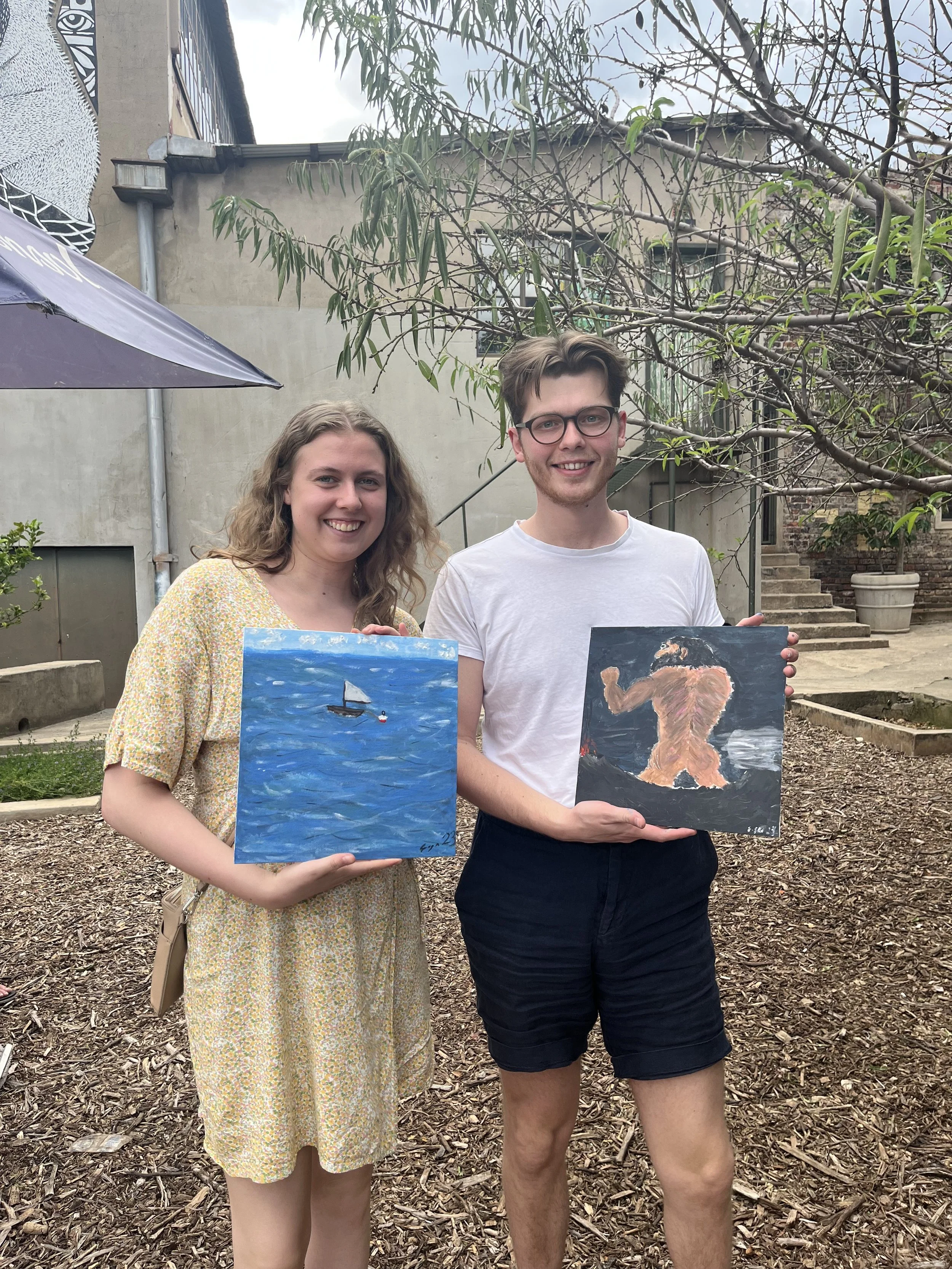 Two young adults, a woman and a man, standing outdoors in front of a building with stairs and trees, holding paintings. The woman has long, curly hair and is wearing a yellow floral dress. The man has short hair, glasses, and is dressed in a white t-