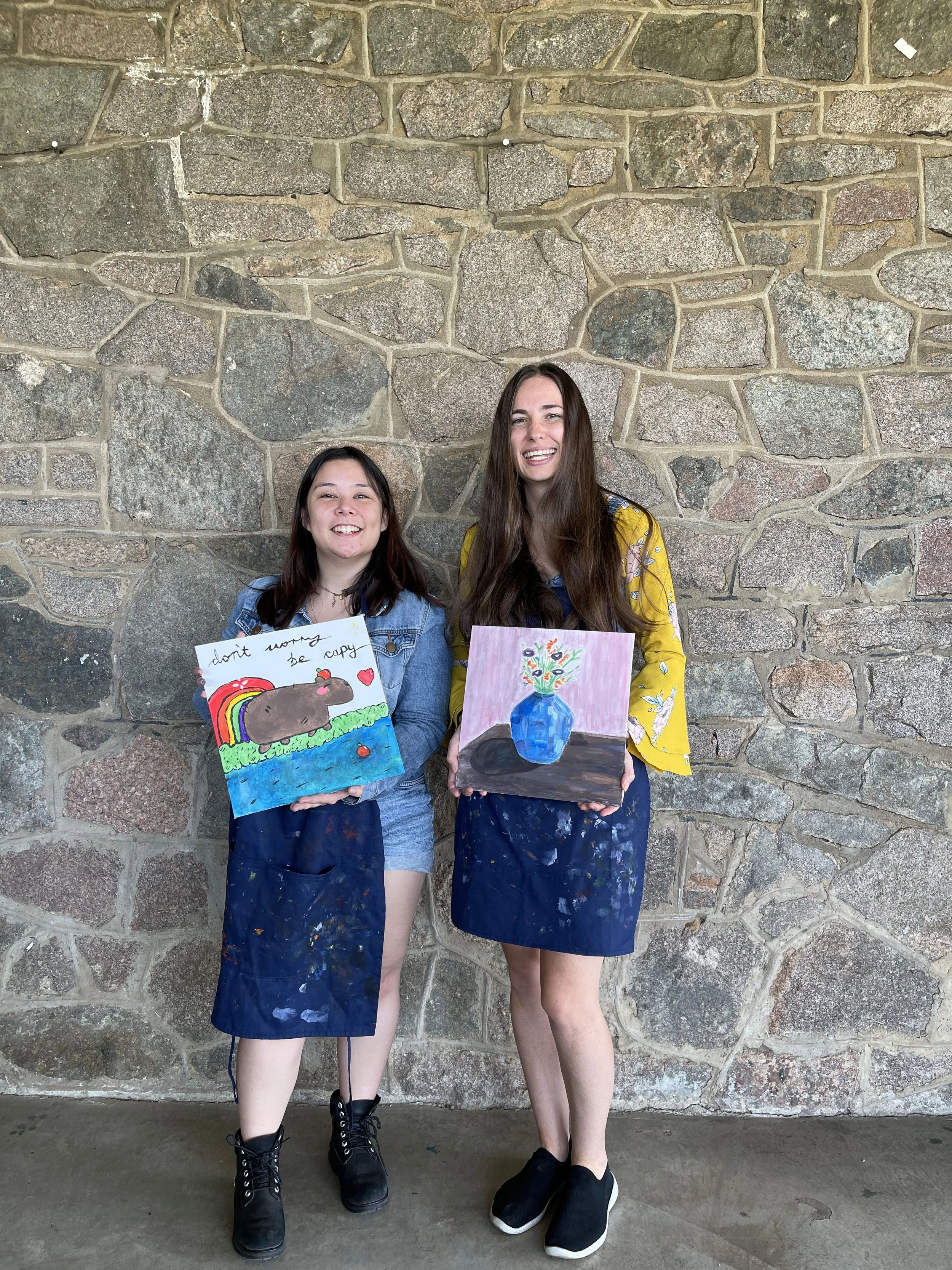 Two young women standing in front of a stone wall, smiling and holding colorful paintings.