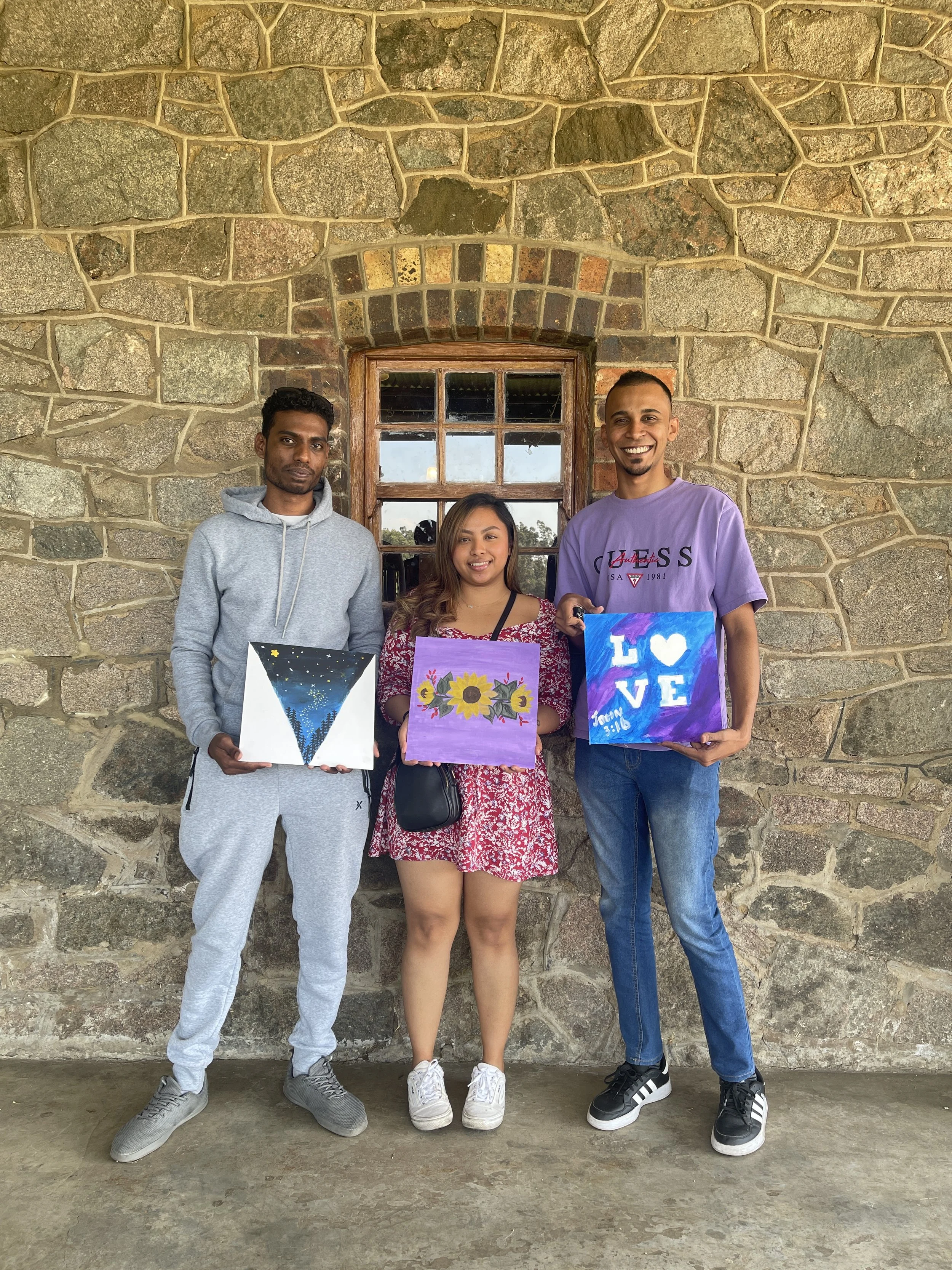 Three young adults standing against a stone wall, each holding a colorful painting, smiling for the camera.
