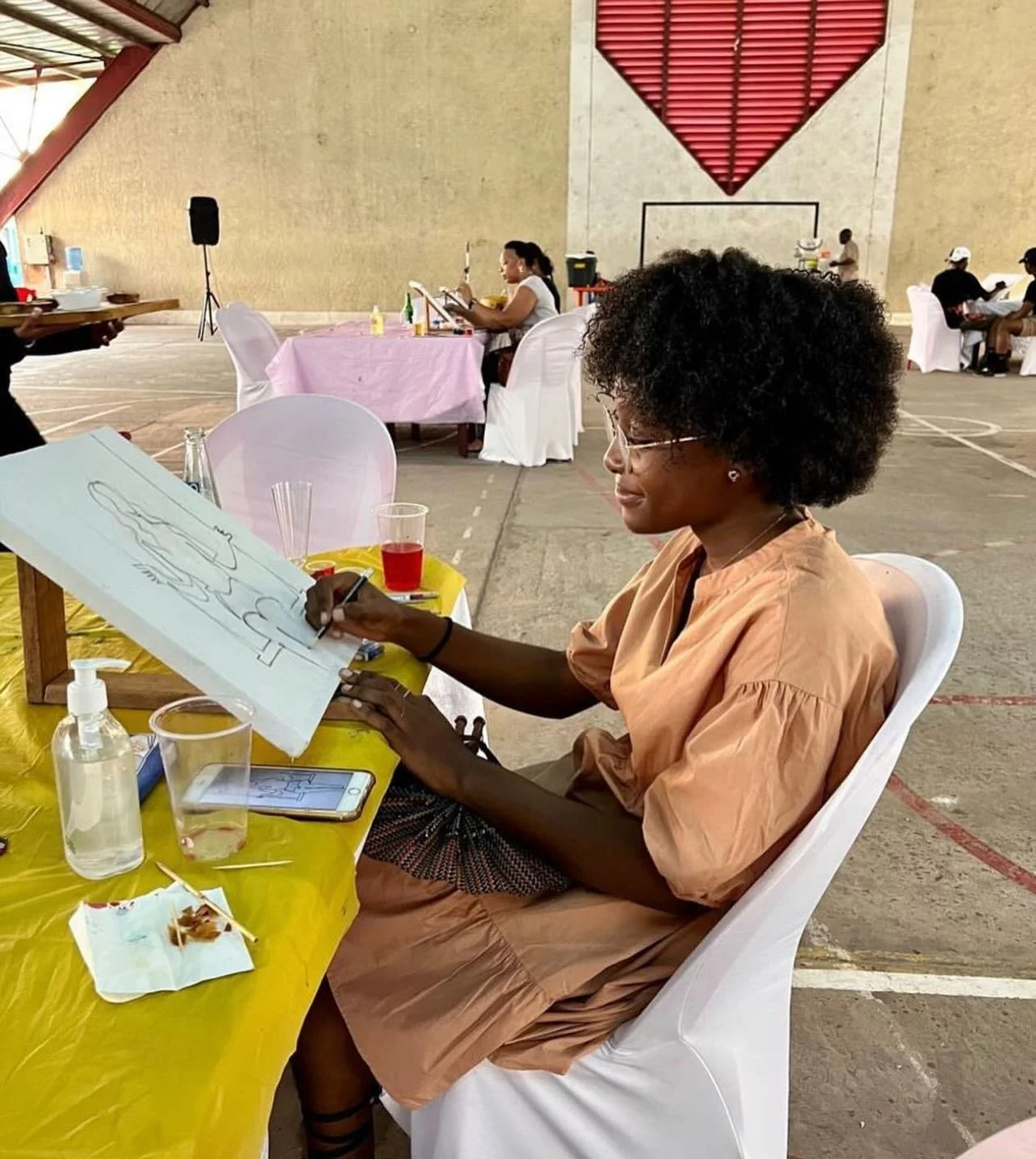 A woman with black curly hair and glasses is drawing on an easel at a table covered with a yellow tablecloth. The table has a smartphone, a drink, a sanitizer bottle, and some papers. Behind her, other people are seated at tables in a large indoor gy
