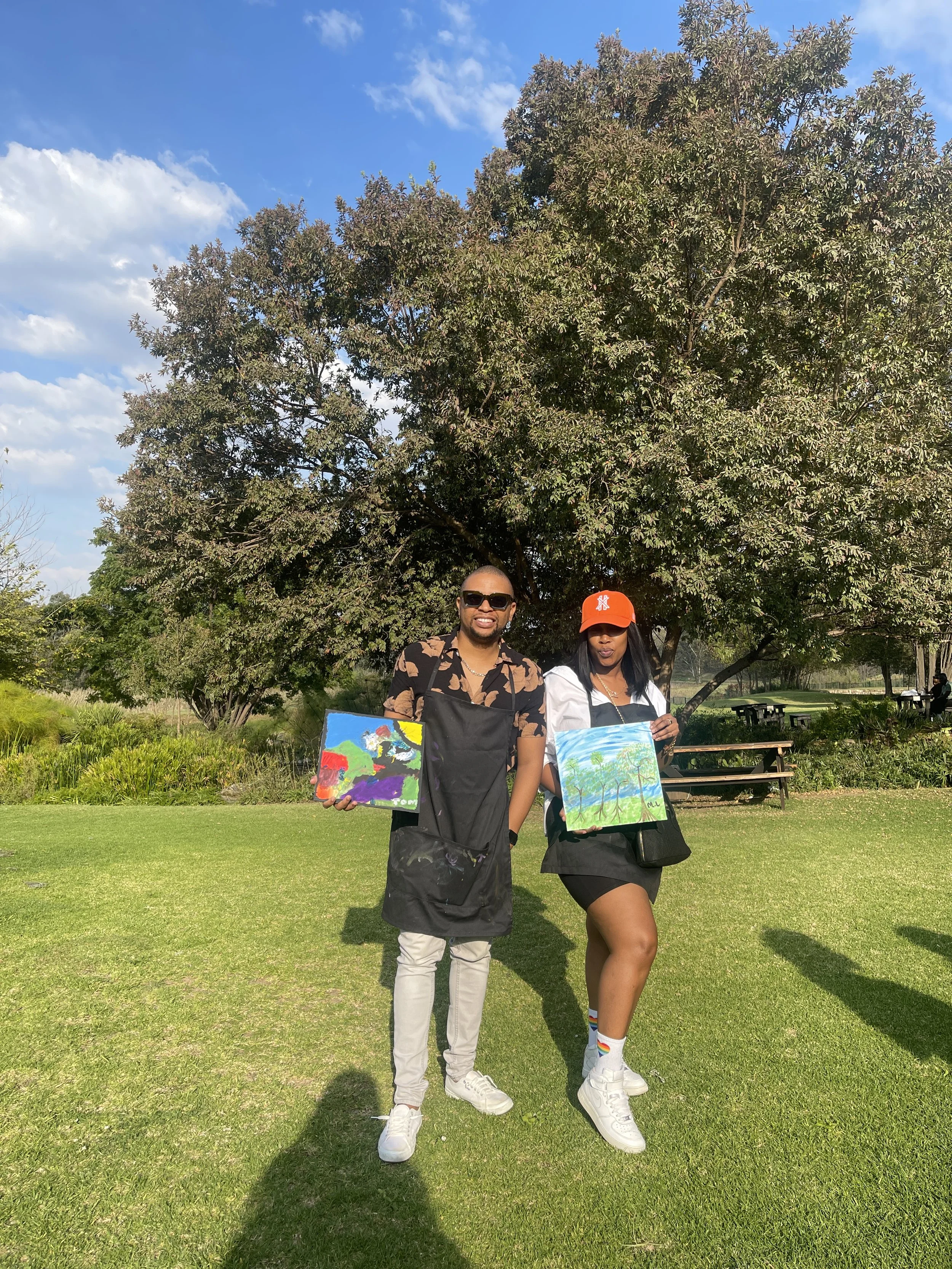 Two people standing on grass holding paintings, with a large tree and blue sky in the background.