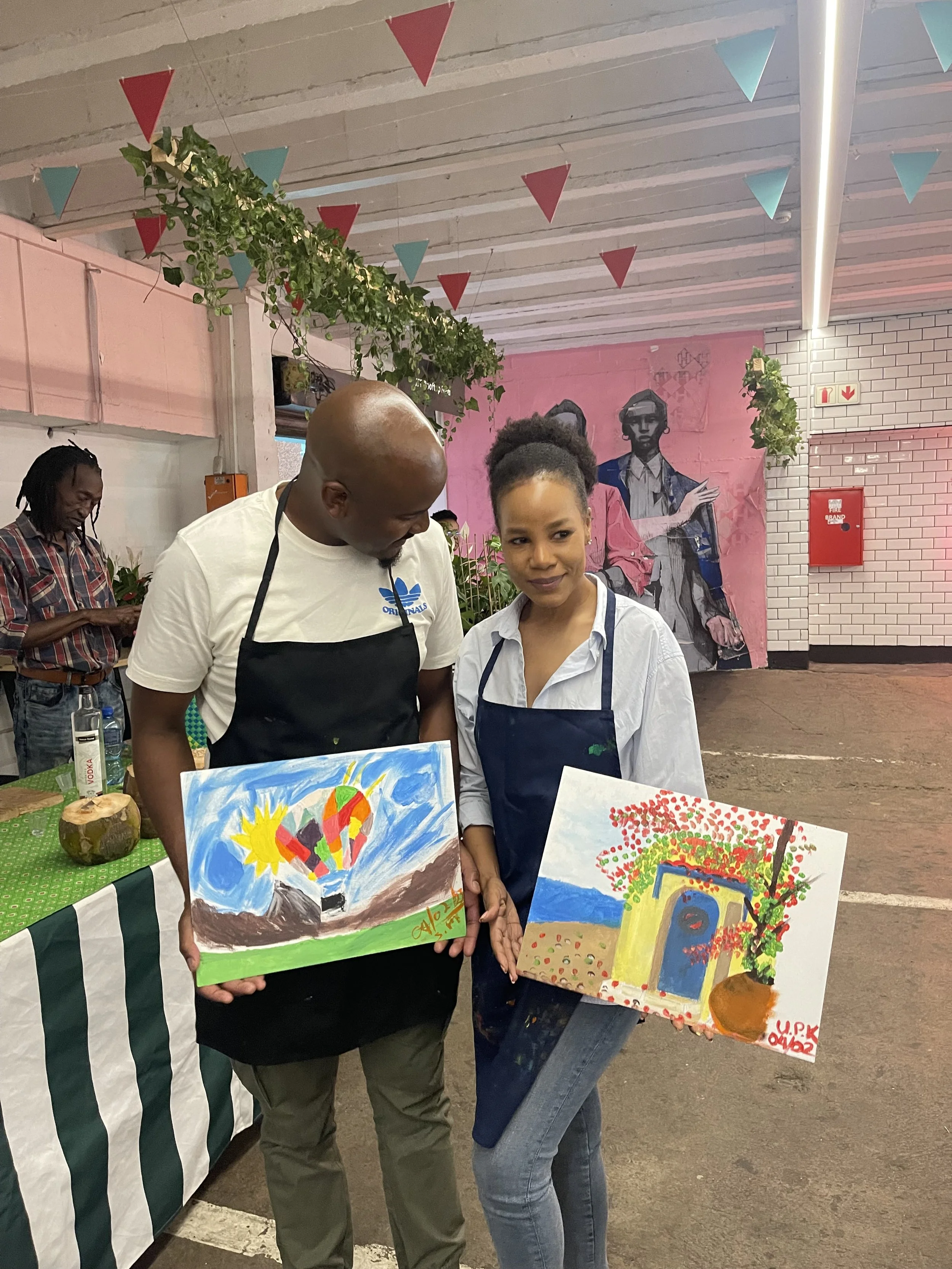Two women are standing together, each holding a colorful painting of trees, a house, and a hot air balloon. One woman wears a gray shirt and apron, and the other wears a white shirt and apron. In the background, a mural of two women is on a pink wall
